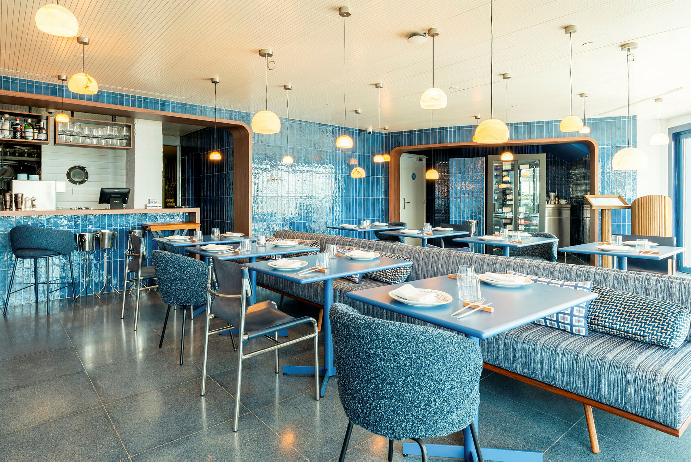 En fargerik restaurantinteriør med blå vegger, bord og stoler. Lyse lamper henger fra taket. (AI-generert tekst)