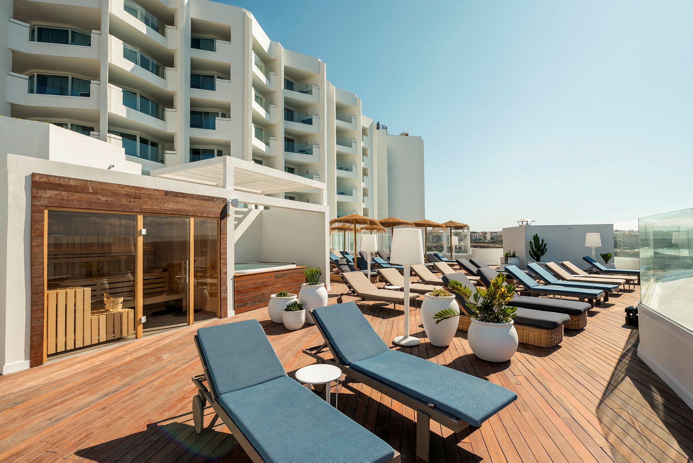 En terrasse med solsenger, parasoller og planter. I bakgrunnen ses et lysmalt hotell. (AI-generert tekst)