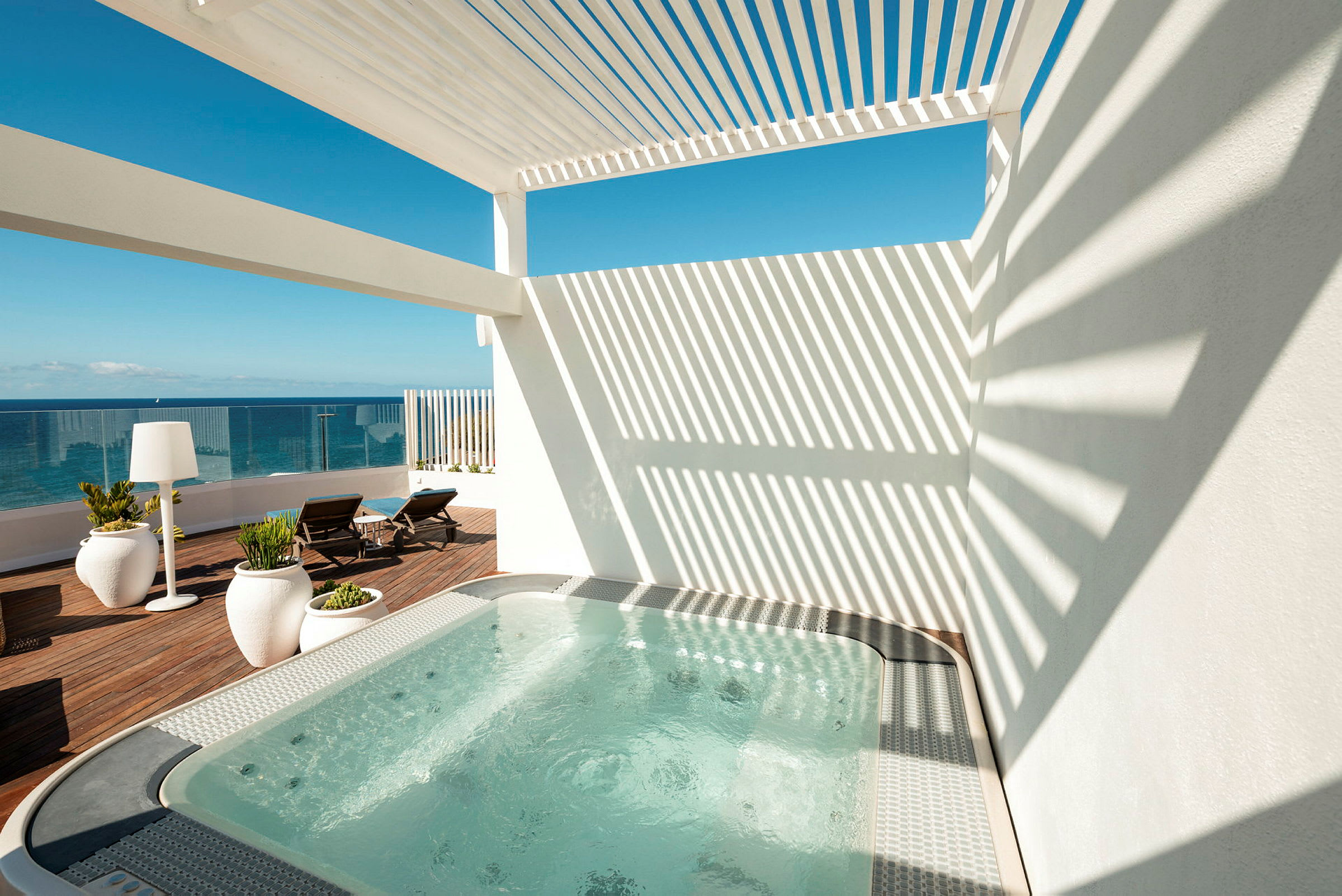 En terrasse med boblebad og utsikt over havet. Terrassen har solsenger, store planter i potter og en pergola som gir skyggefulle områder. (AI-generert tekst)