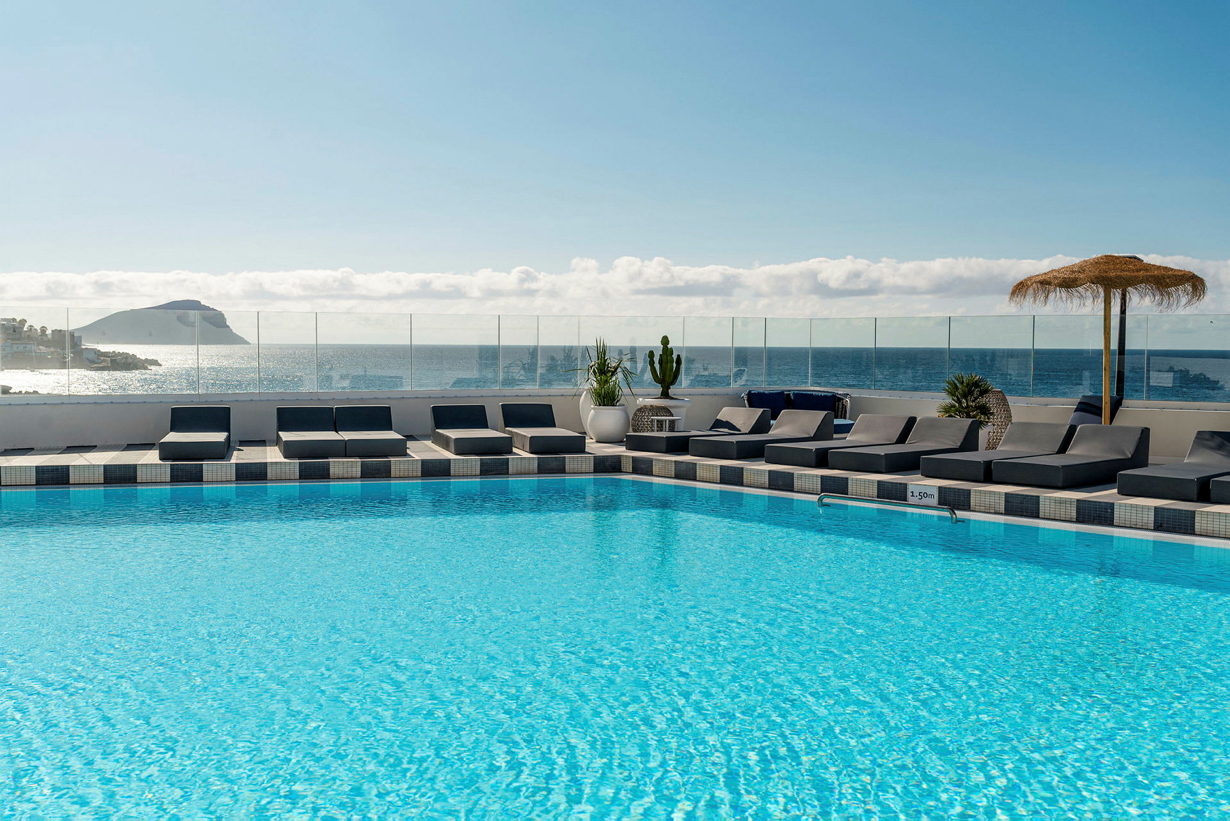 Et basseng på et hotell med solsenger rundt samt utsikt over havet og en øy i bakgrunnen. (AI-generert tekst)