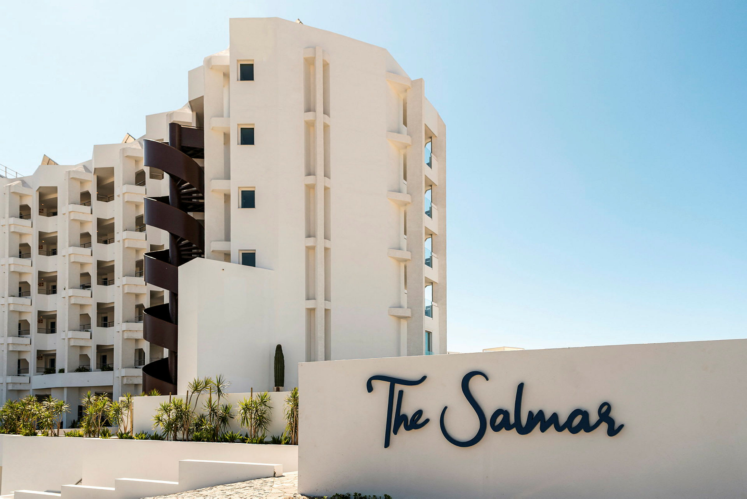 En moderne bygning med balkonger og en spiraltrapp ved siden av et skilt med teksten "The Salmar". Foran bygningen er det beplantning med palmer. (AI-generert tekst)