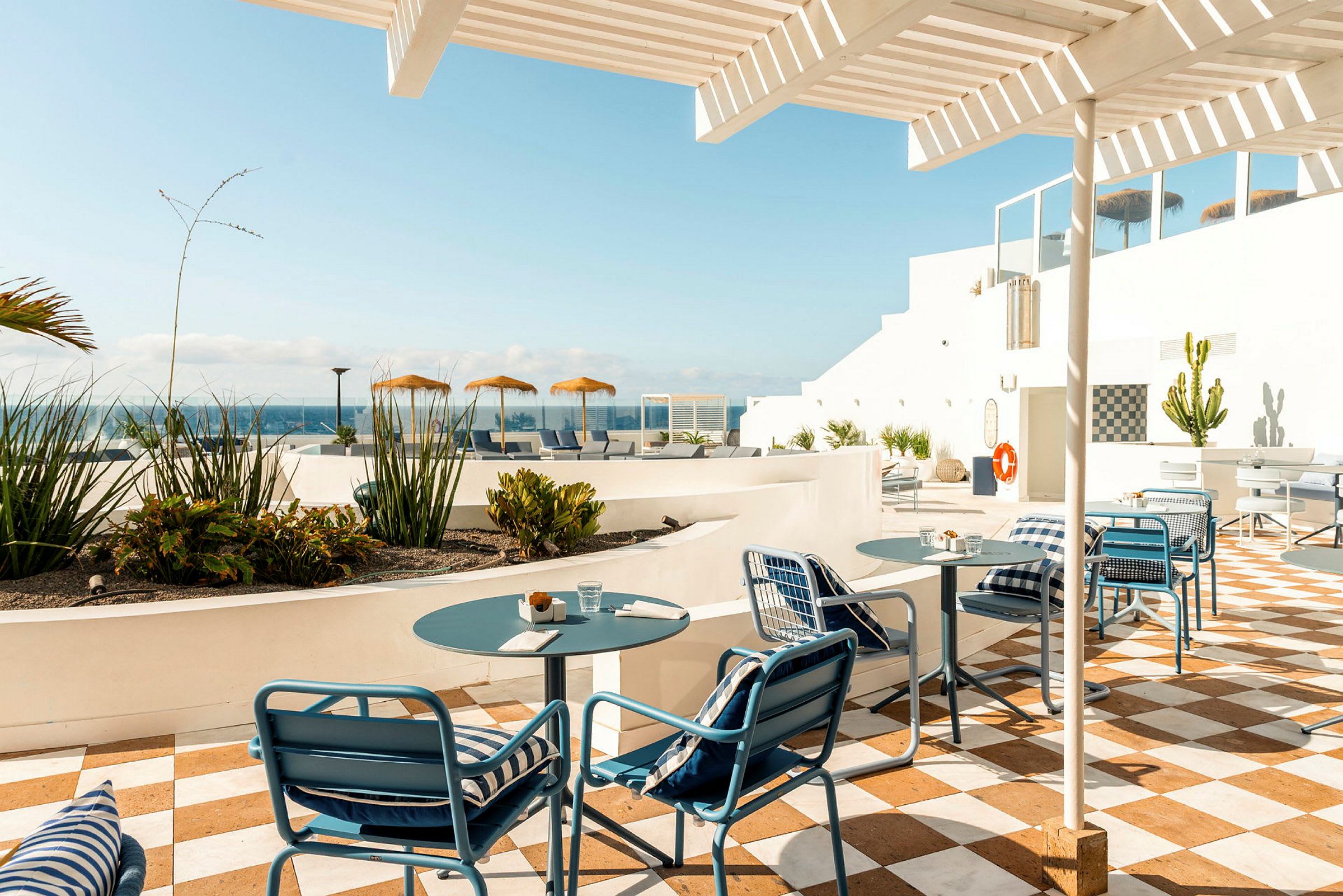 Utendørs serveringsområde med bord og stoler under en pergola, omgitt av vegetasjon og med utsikt over havet. (AI-generert tekst)