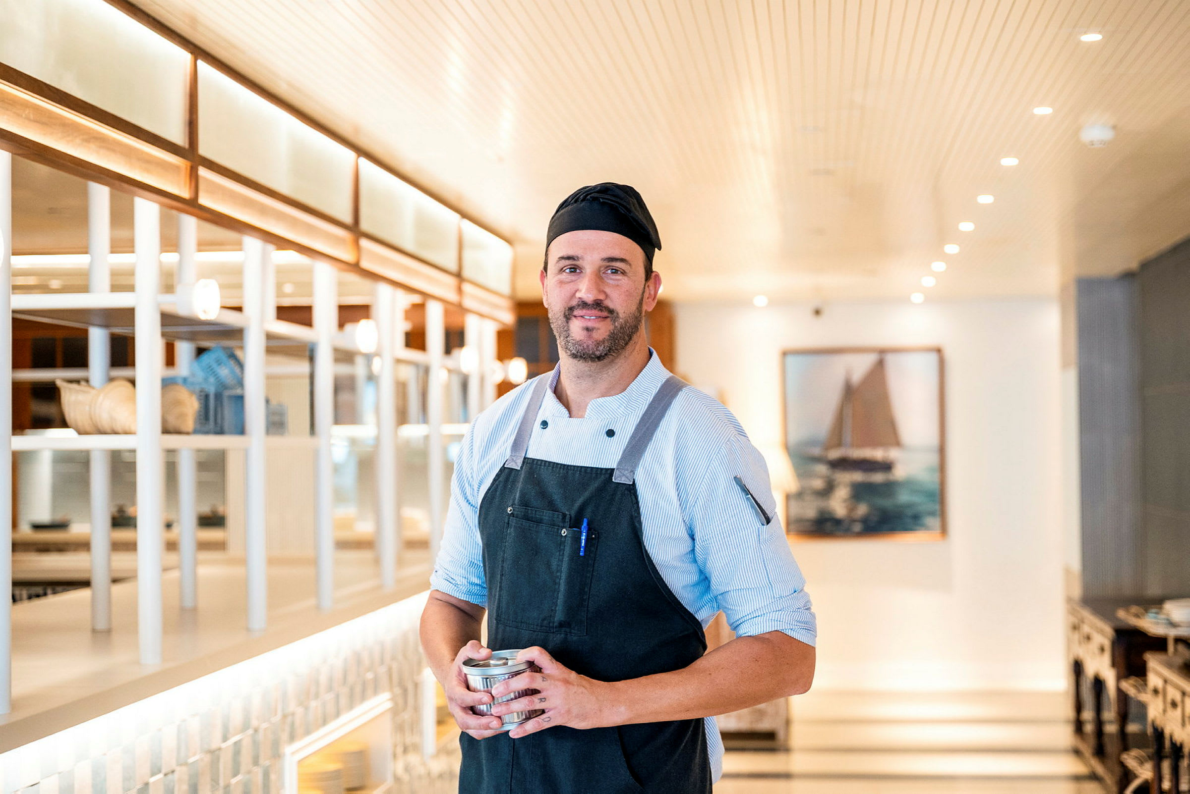 En kokk i arbeidsklær står i et lyst restaurantmiljø med moderne innredning. (AI-generert tekst)