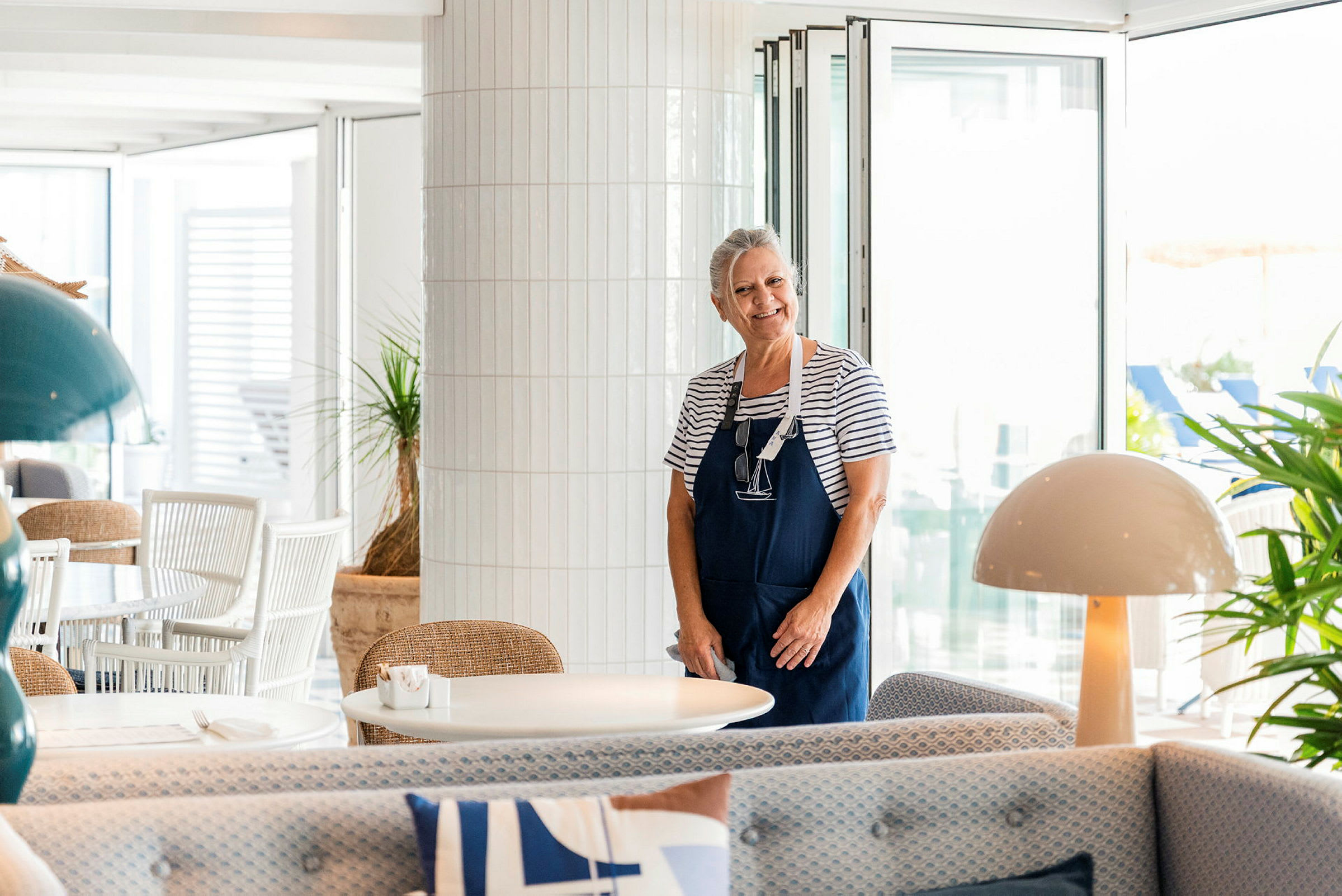 En kvinne står i en lys og moderne restaurant, kledd i stripet genser og forkle. (AI-generert tekst)
