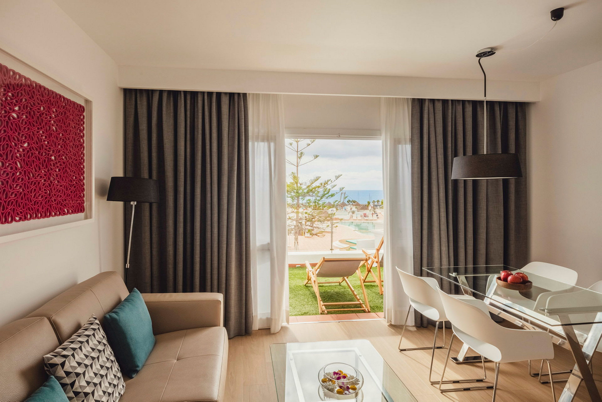 En moderne stue med en beige sofa og spisebord med hvite stoler. Rommet har store vinduer med gardiner og utsikt mot en grønn uteplass med solsenger og et basseng. (AI-generert tekst)