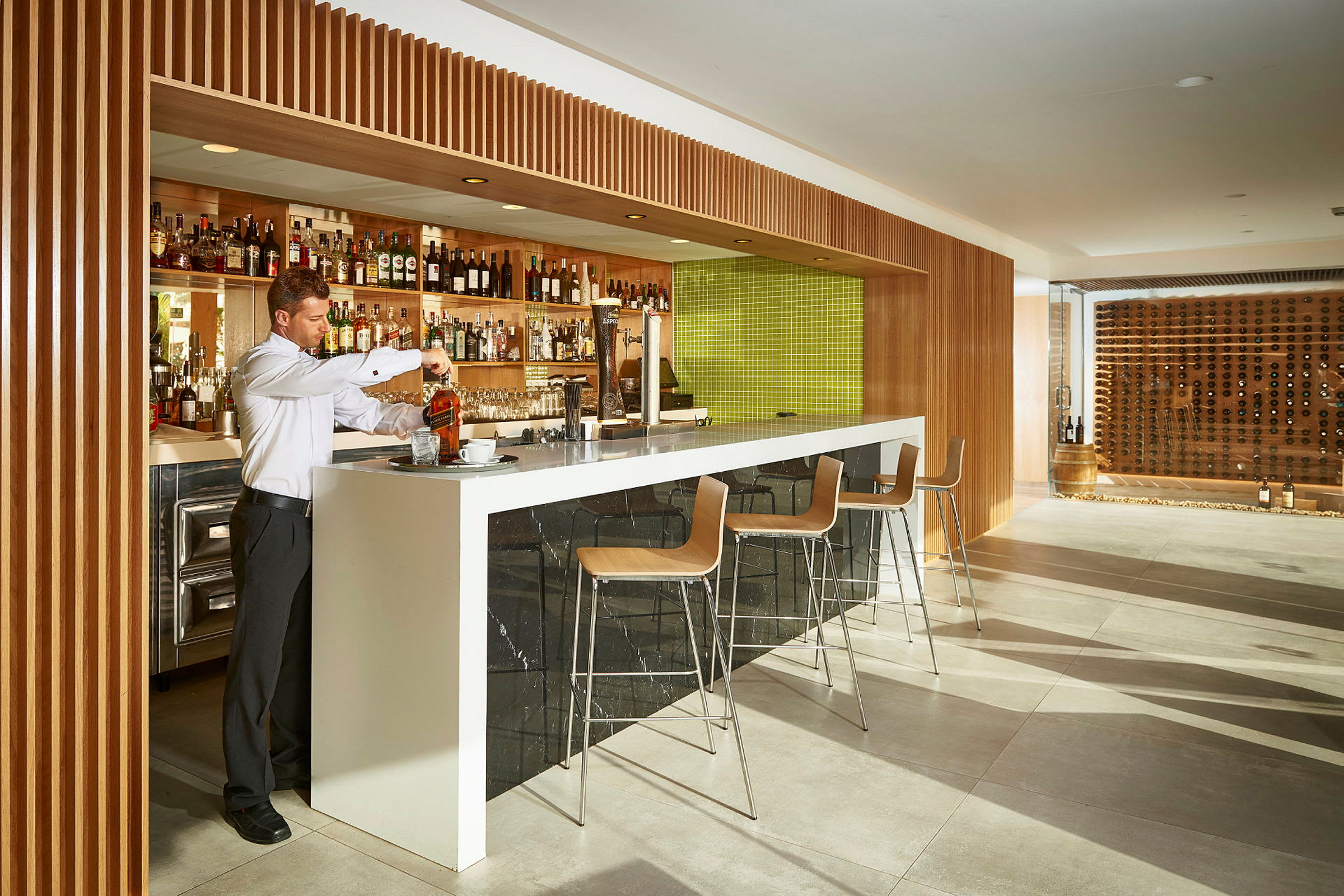 En bartender forbereder en drink ved en moderne bar med trepaneler og en grønn flislagt bakgrunn. Barstoler står klare ved disken. (AI-generert tekst)