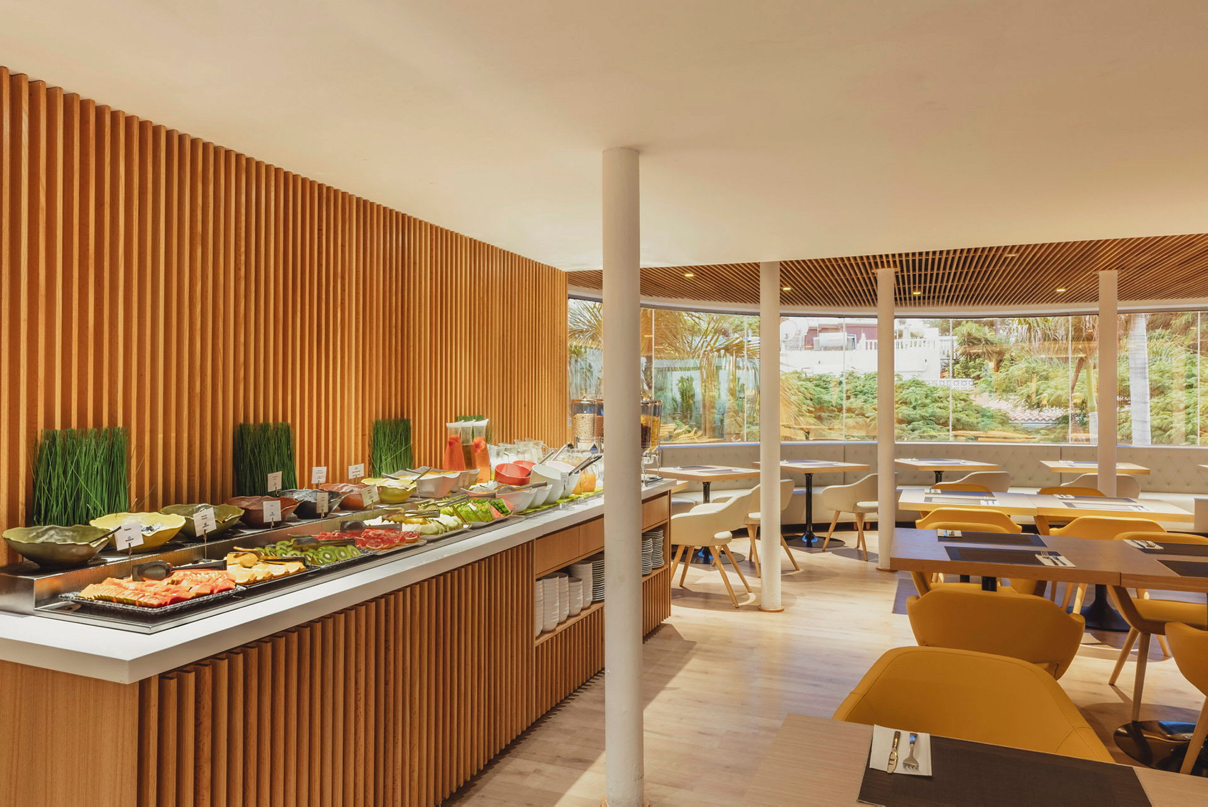 Bilde av en restaurant med en buffé med ulike matretter og drikkevarer, omgitt av sitteplasser med gulaktige stoler og store vinduer som viser en grønn hage utenfor. (AI-generert tekst)