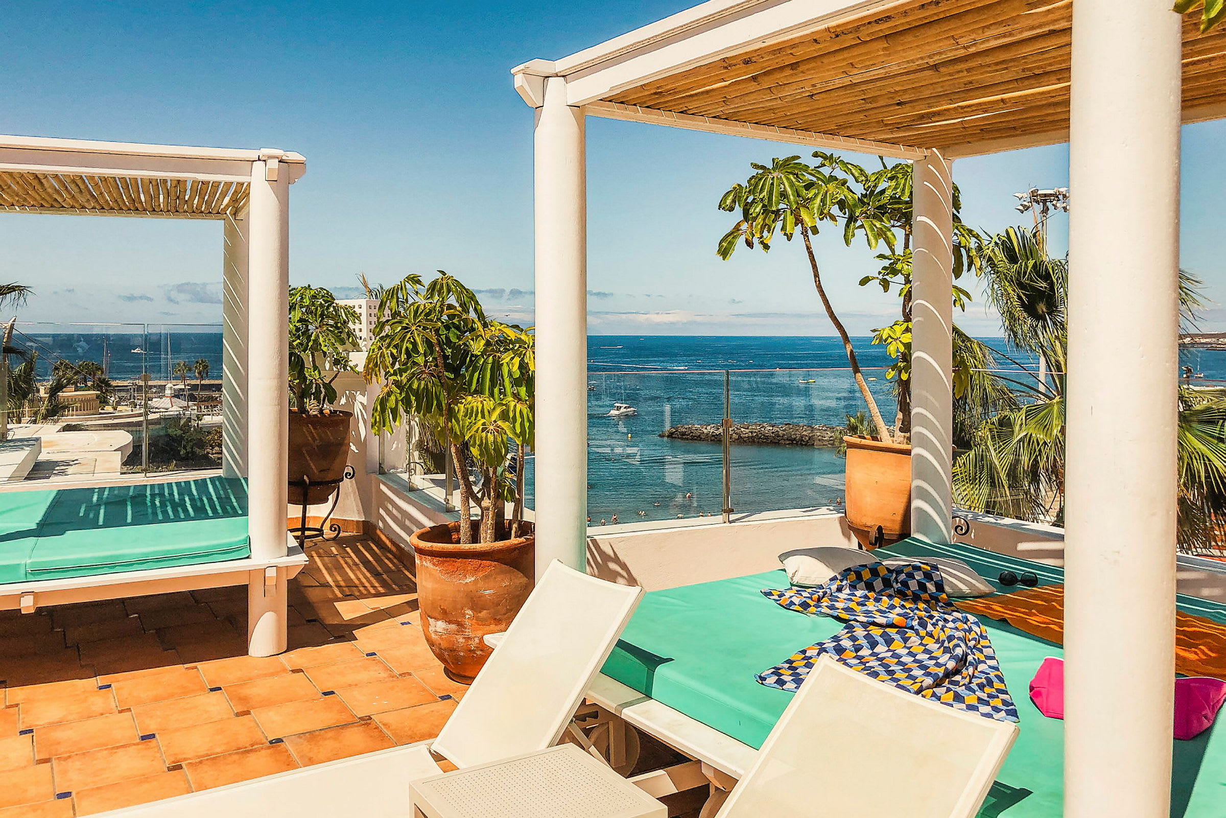 En solrik terrasse med utsikt over havet. En dagseng med turkis madrass og fargerike stoffer står under en pergola. Rundt terrassen er det potteplanter og palmer. (AI-generert tekst)