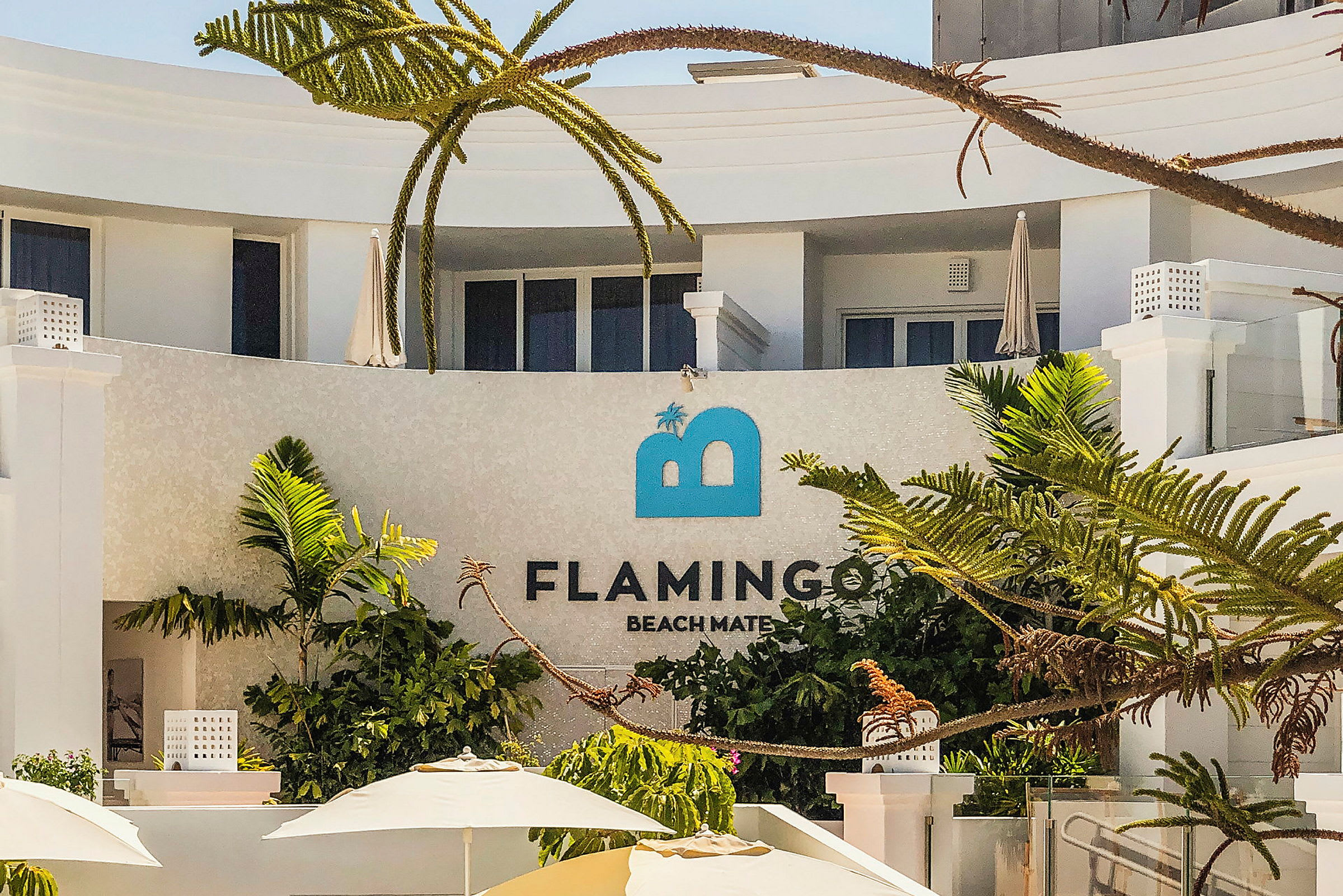 En moderne bygning med et skilt hvor det står Flamingo Beach Mate, omgitt av palmer og parasoller. (AI-generert tekst)