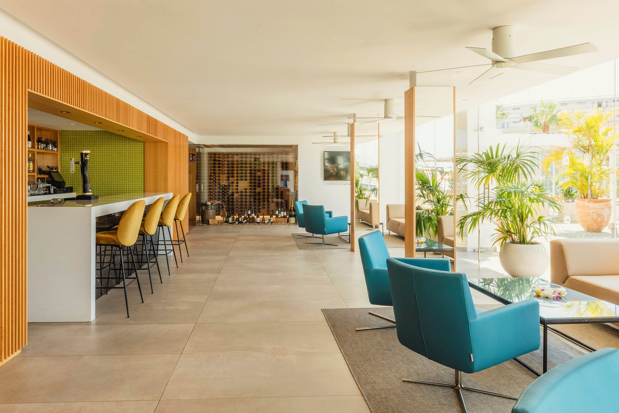 Et moderne innredet caféområde med en bardisk og grønne flisvegger, ryggstoler med gullfargede trekk, samt komfortable sitteplasser med blå og beige lenestoler. Grøntområder og glassvegger ses i bakgrunnen. (AI-generert tekst)