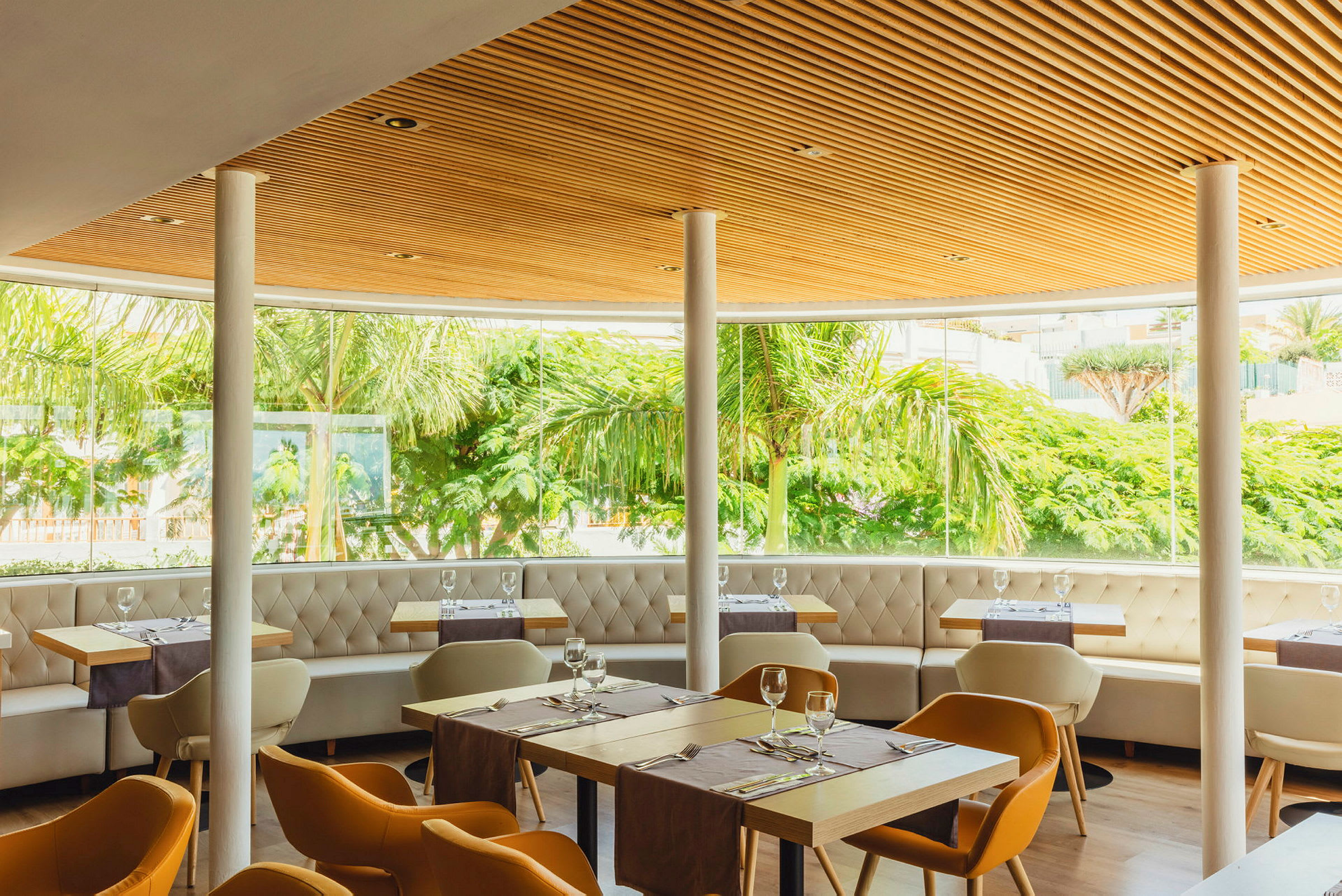 En moderne restaurantinteriør med store vinduer som gir utsikt over grøntområder. Møbler inkluderer stoler og bord, samt sofaer langs vinduene. (AI-generert tekst)