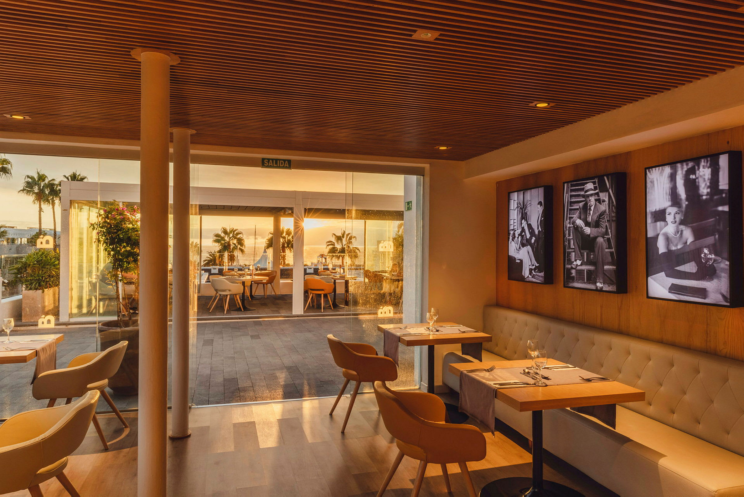 En innendørs del av en restaurant med bord og stoler, samt glasspartier som viser utsikt mot en utendørsterrasse med bord og palmer i solnedgang. (AI-generert tekst)