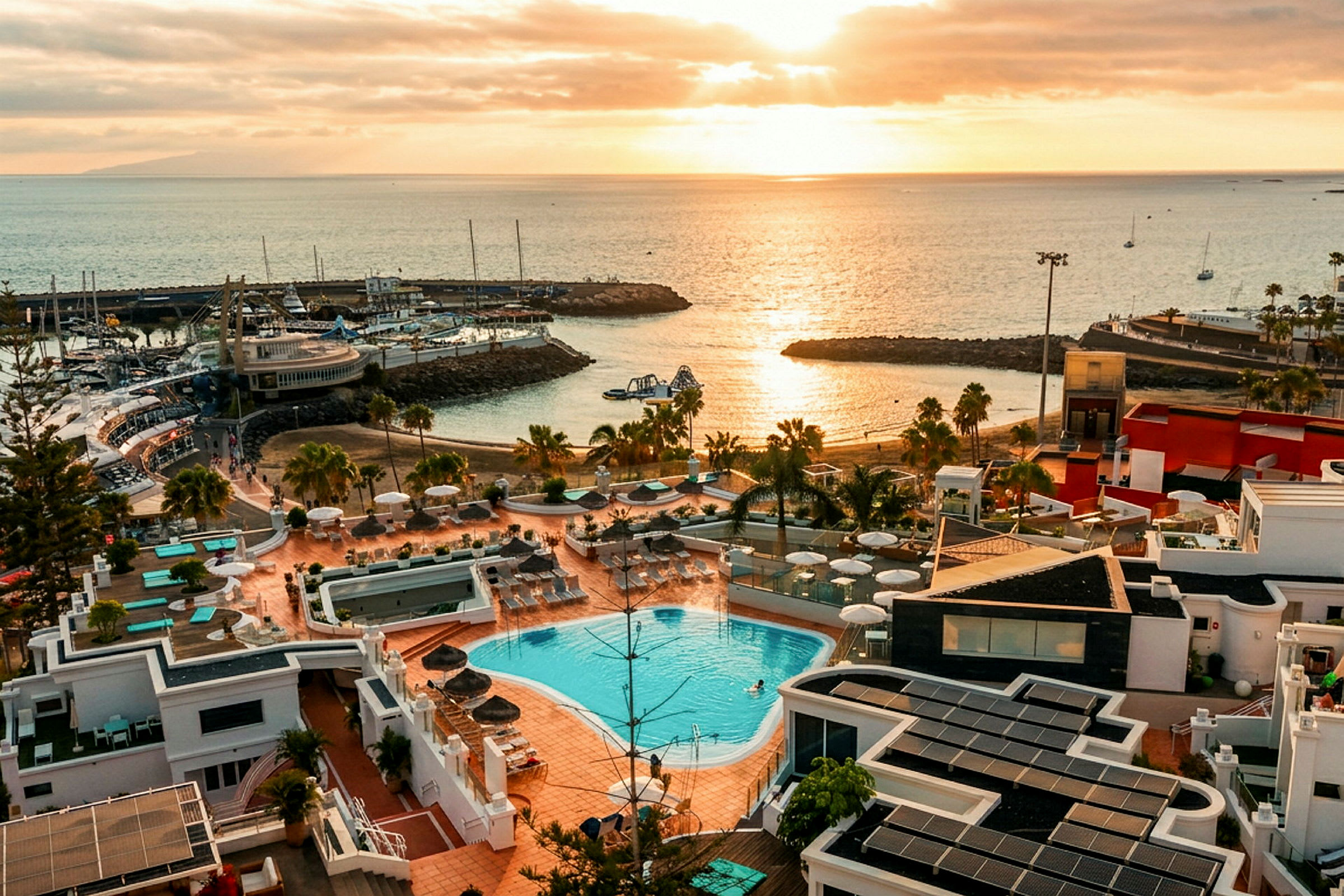 Utsikt over et kystområde ved solnedgang med et anlegg som har basseng og solterrasse i forgrunnen samt havet og småbåter i bakgrunnen. (AI-generert tekst)