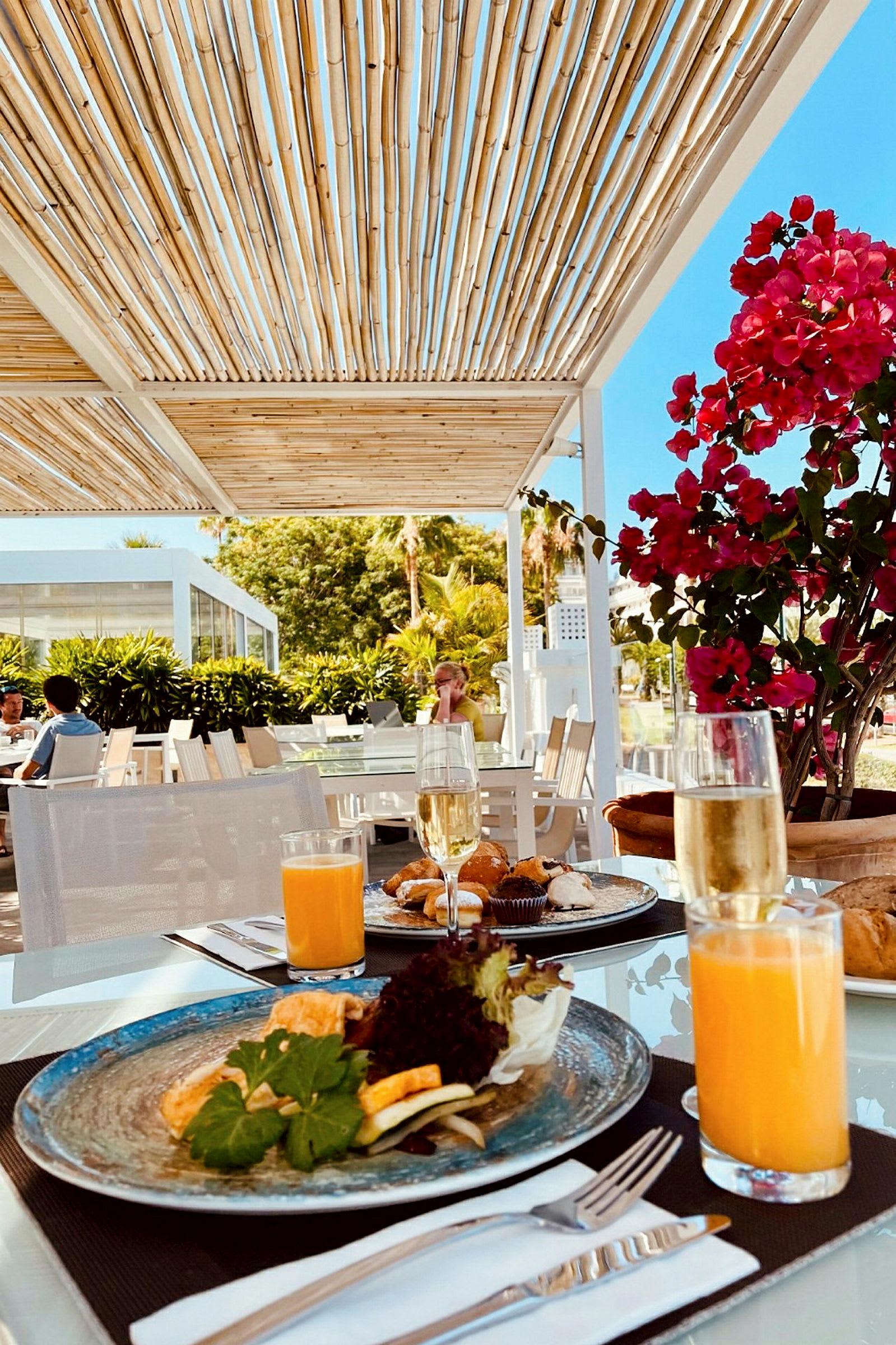 Et bord er dekket med tallerkener, glass med juice og champagne, samt bestikk. I bakgrunnen er en pergola, planter og mennesker som sitter ved andre bord. (AI-generert tekst)
