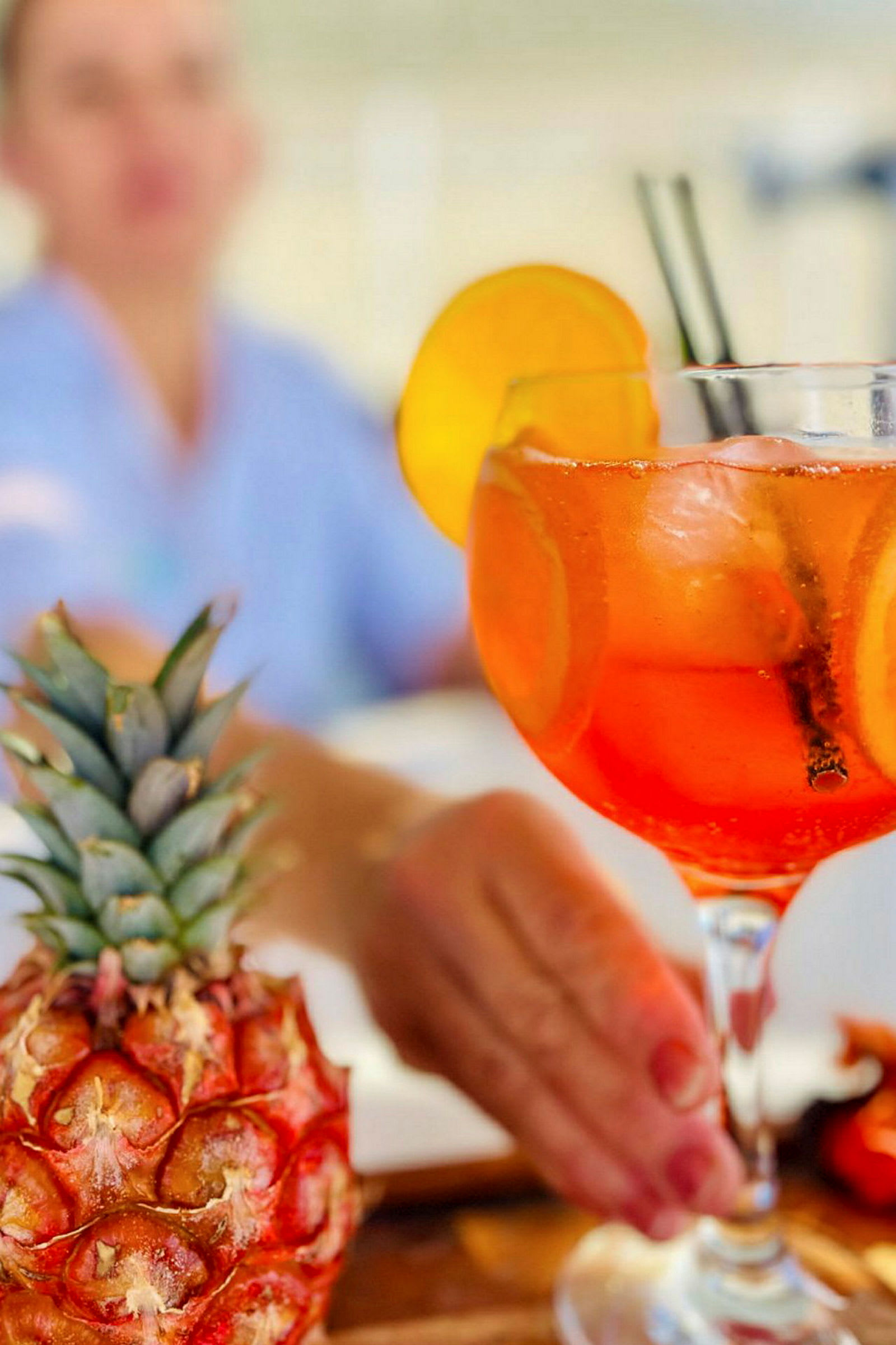 Et nærbilde av en drink i et glass med appelsinskiver og isbiter. En ananas er synlig i forgrunnen. (AI-generert tekst)