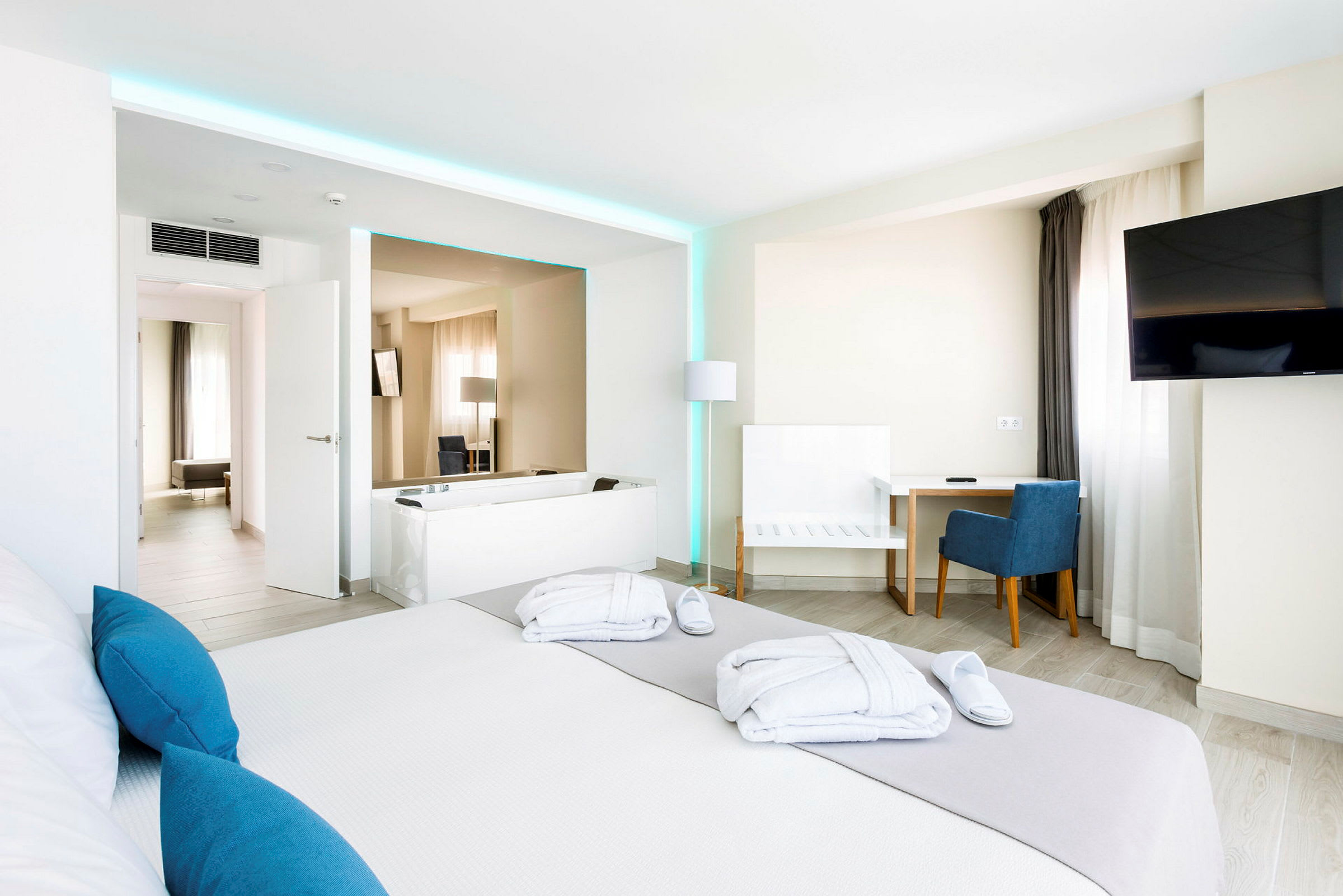Et moderne hotellrom med en stor seng, skrivebord og stol, en TV på veggen, samt et badekar. Lyse vegger og interiør med blå aksenter. (AI-generert tekst)