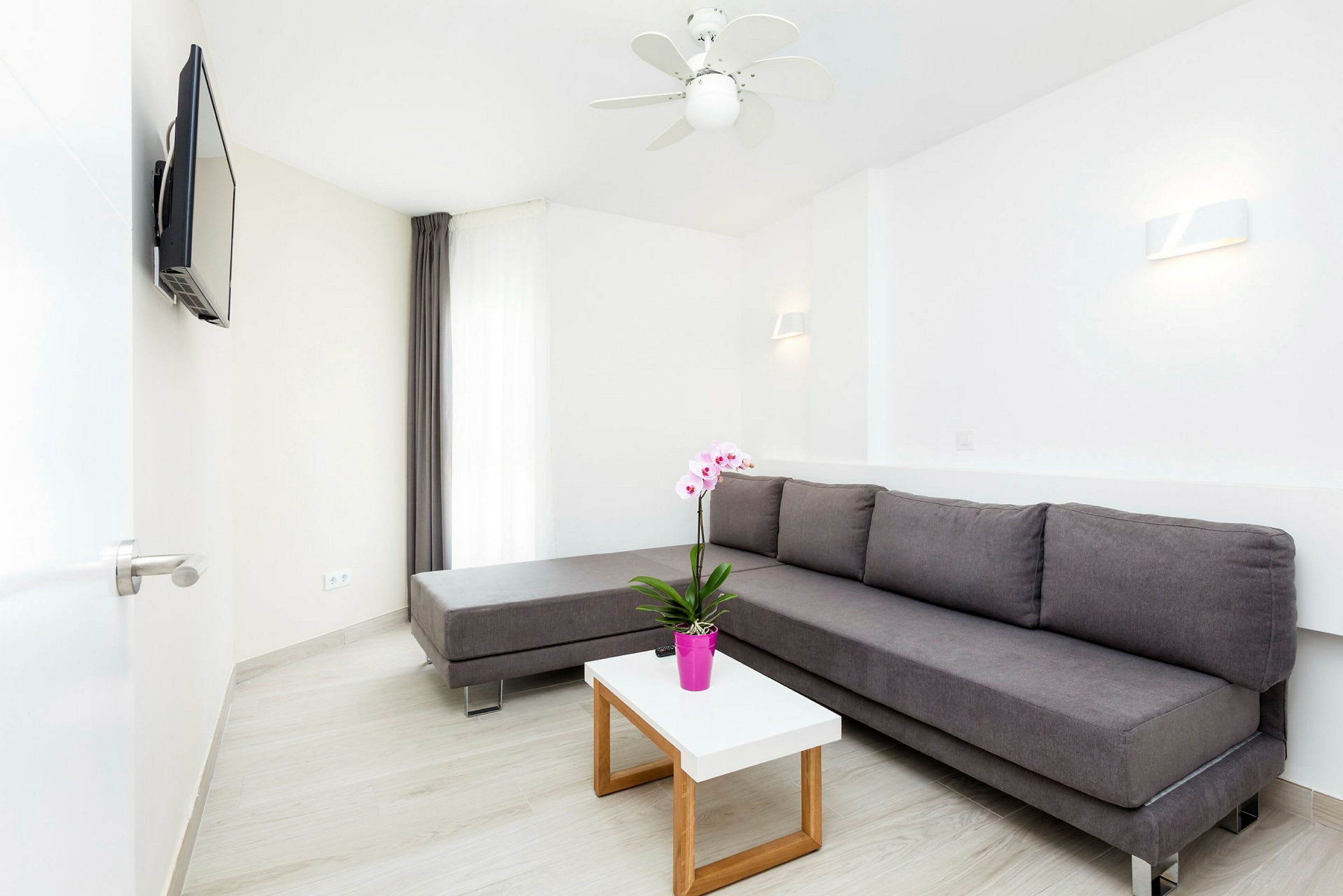 En moderne lounge med en grå sofa, hvite vegger og et lite bord med en rosa blomsterpotte. (AI-generert tekst)