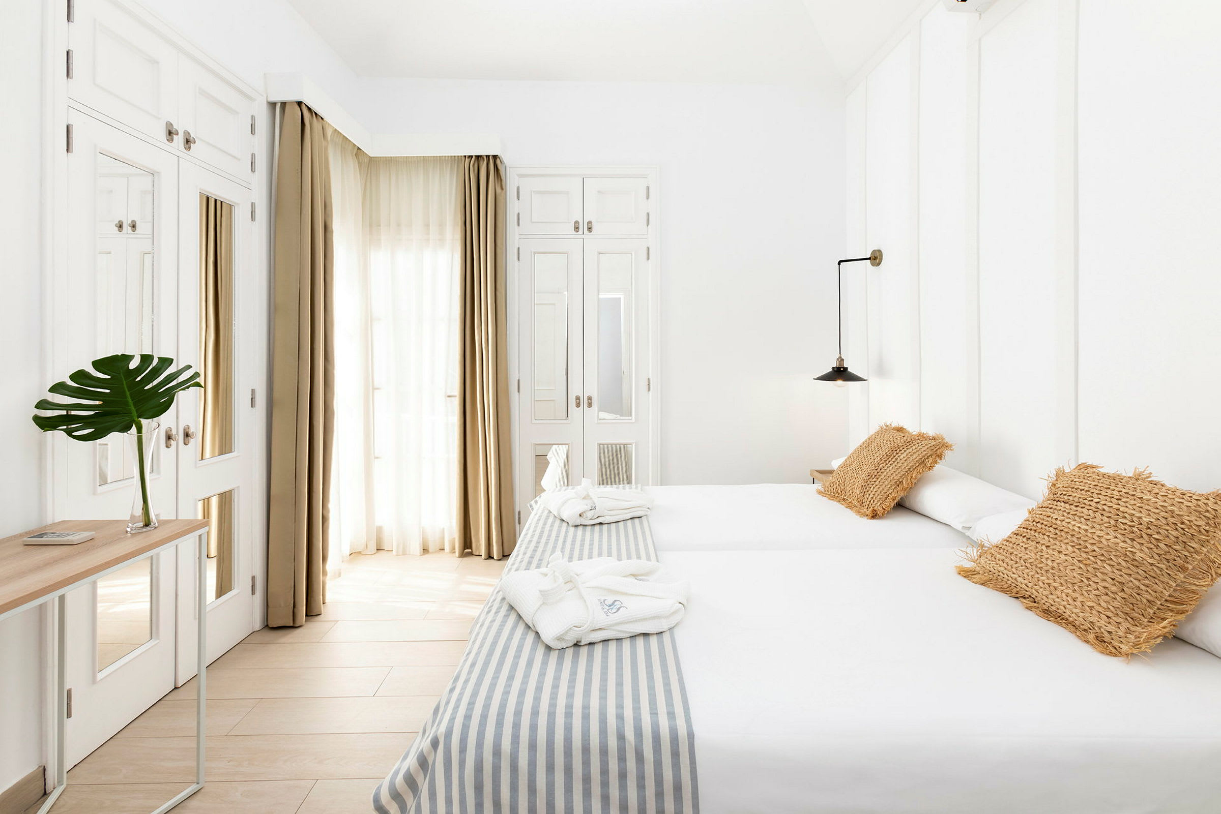 Et lyst hotellrom med to enkeltsenger, beige puter og et bord med en grønn plante i en glassvase. (AI-generert tekst)