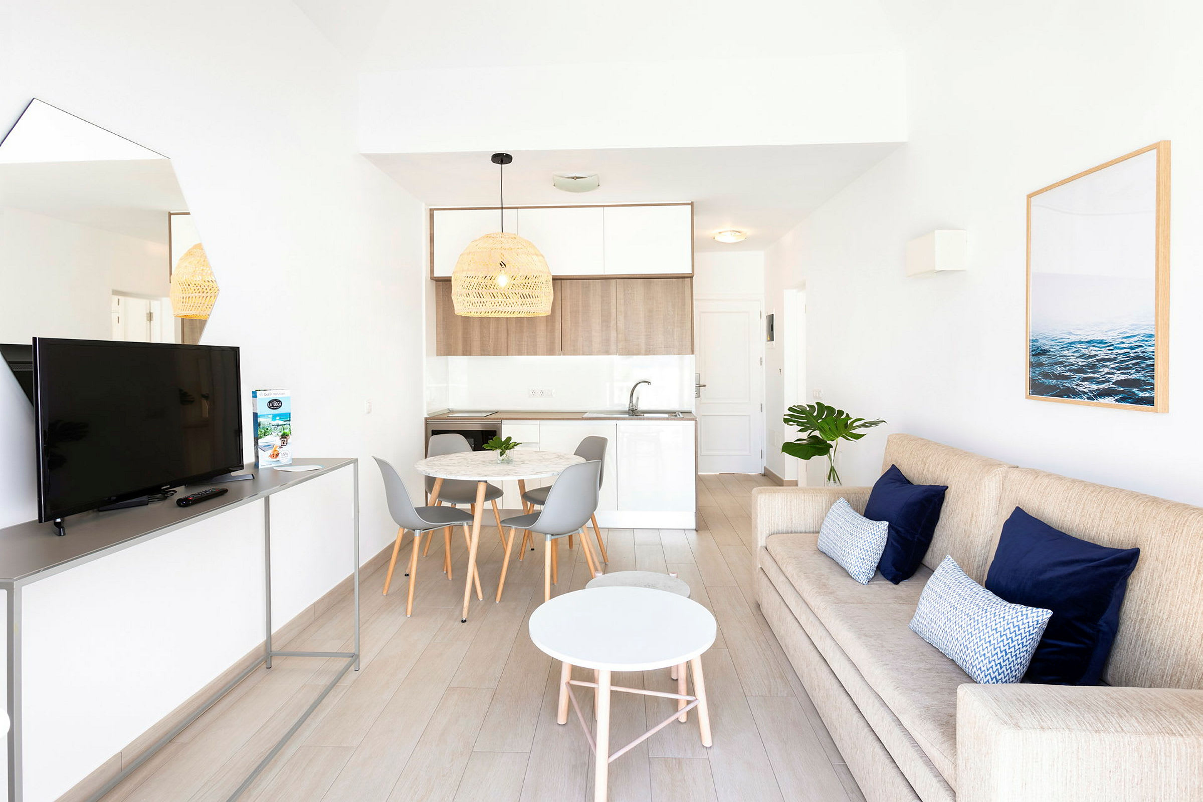 En moderne stue med en beige sofa, blå puter, et hvitt rundt salongbord og en TV på stativ. Ved siden av er det et spiseområde med et rundt bord og grå stoler samt et kjøkken i lyse farger. (AI-generert tekst)