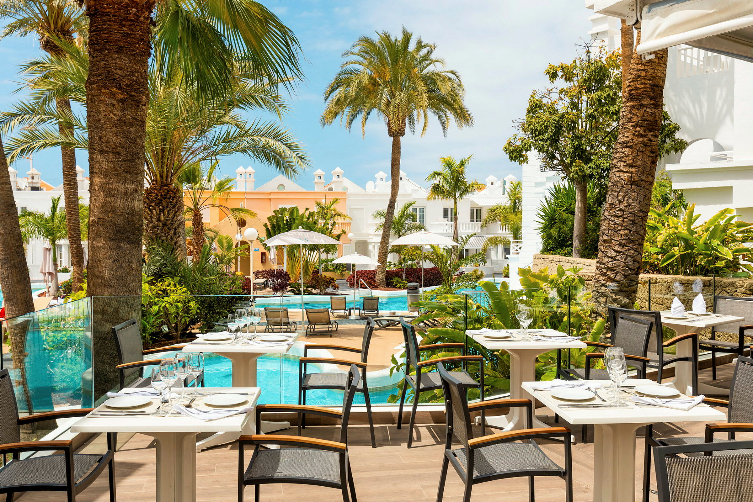 Bilde av en restaurantterrasse med utsikt over et basseng omgitt av palmer og lyse bygninger. (AI-generert tekst)