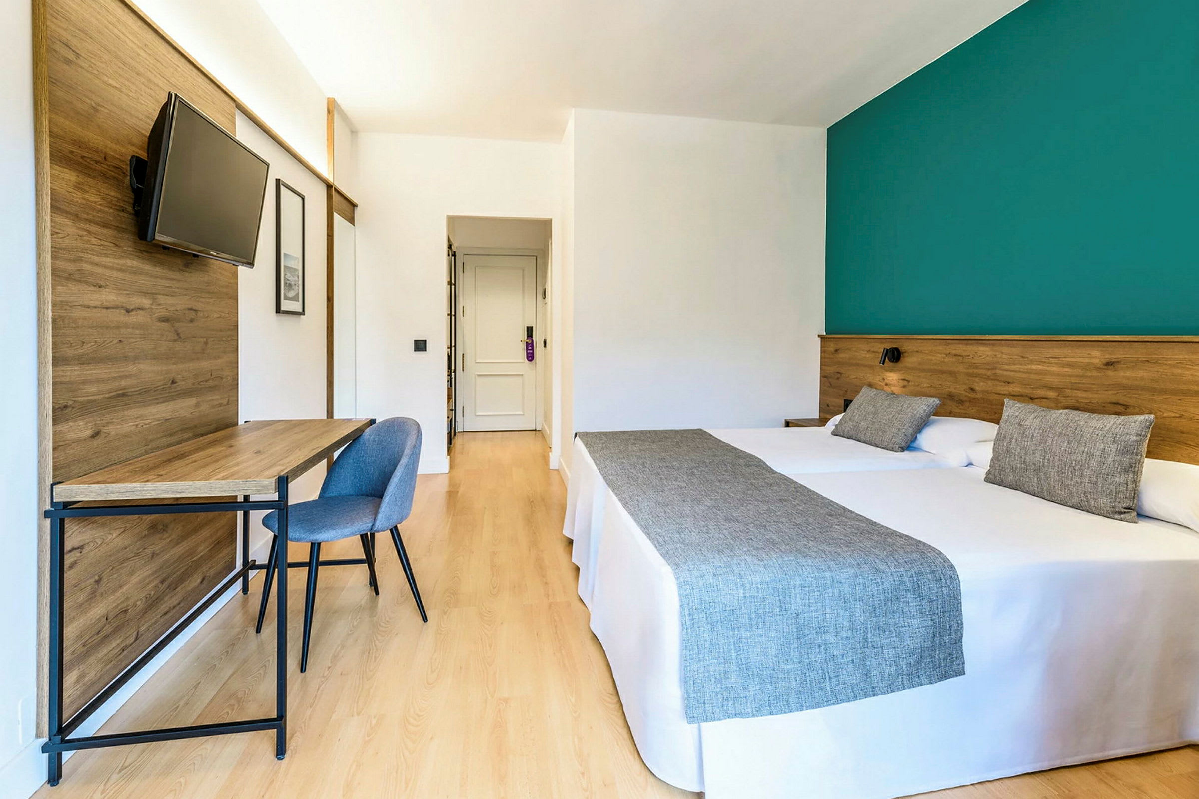 Et hotellrom med to senger, et skrivebord med stol og en flatskjerm-TV på en trefinér. (AI-generert tekst)