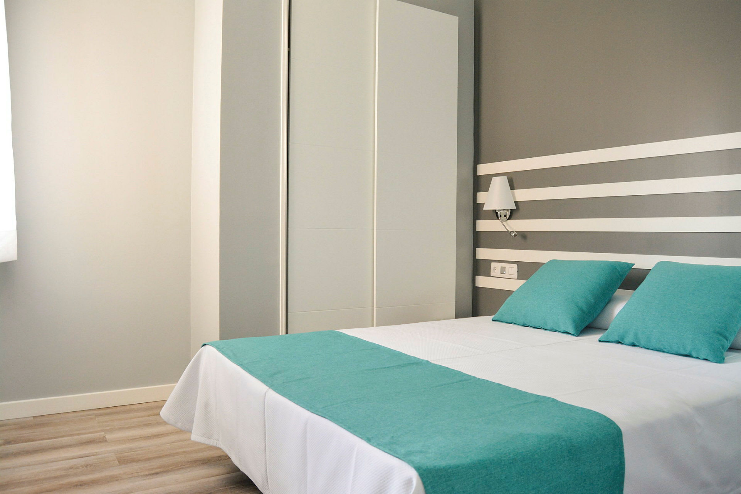 Hotellrom med en dobbeltseng oppredt med hvite laken og turkise puter samt sengeteppet. (AI-generert tekst)