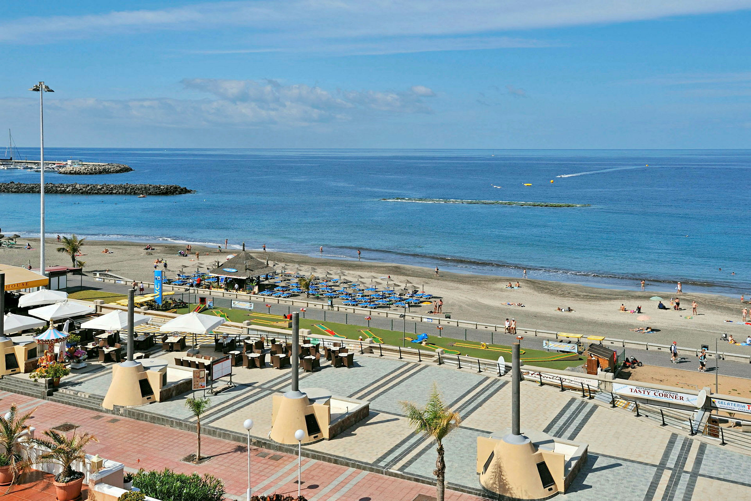 Strand med parasoller, solsenger og folk som soler seg, samt hav og brygger i bakgrunnen. (AI-generert tekst)