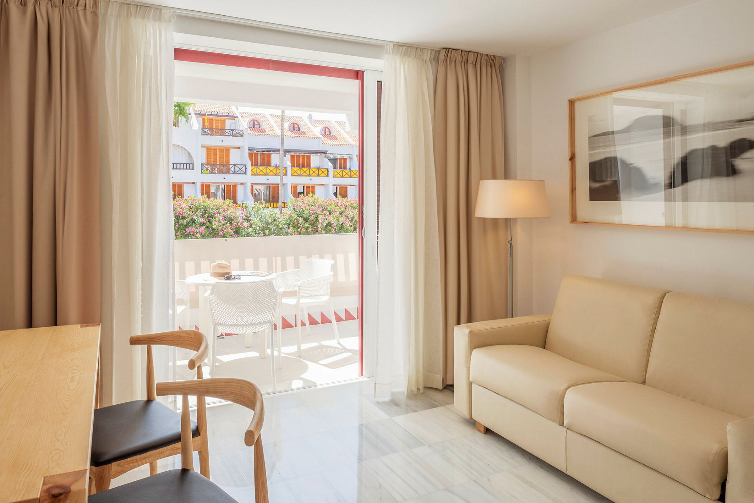 Et romslig og lyst oppholdsrom med beige sofa, tredestoler og et lite spisebord. Store glassdører fører ut til en terrasse med utemøbler, hvor man kan se blomstrende busker og fargerike bygninger. På veggen finnes et innrammet bilde og en gulvlampe. (AI-generert tekst)