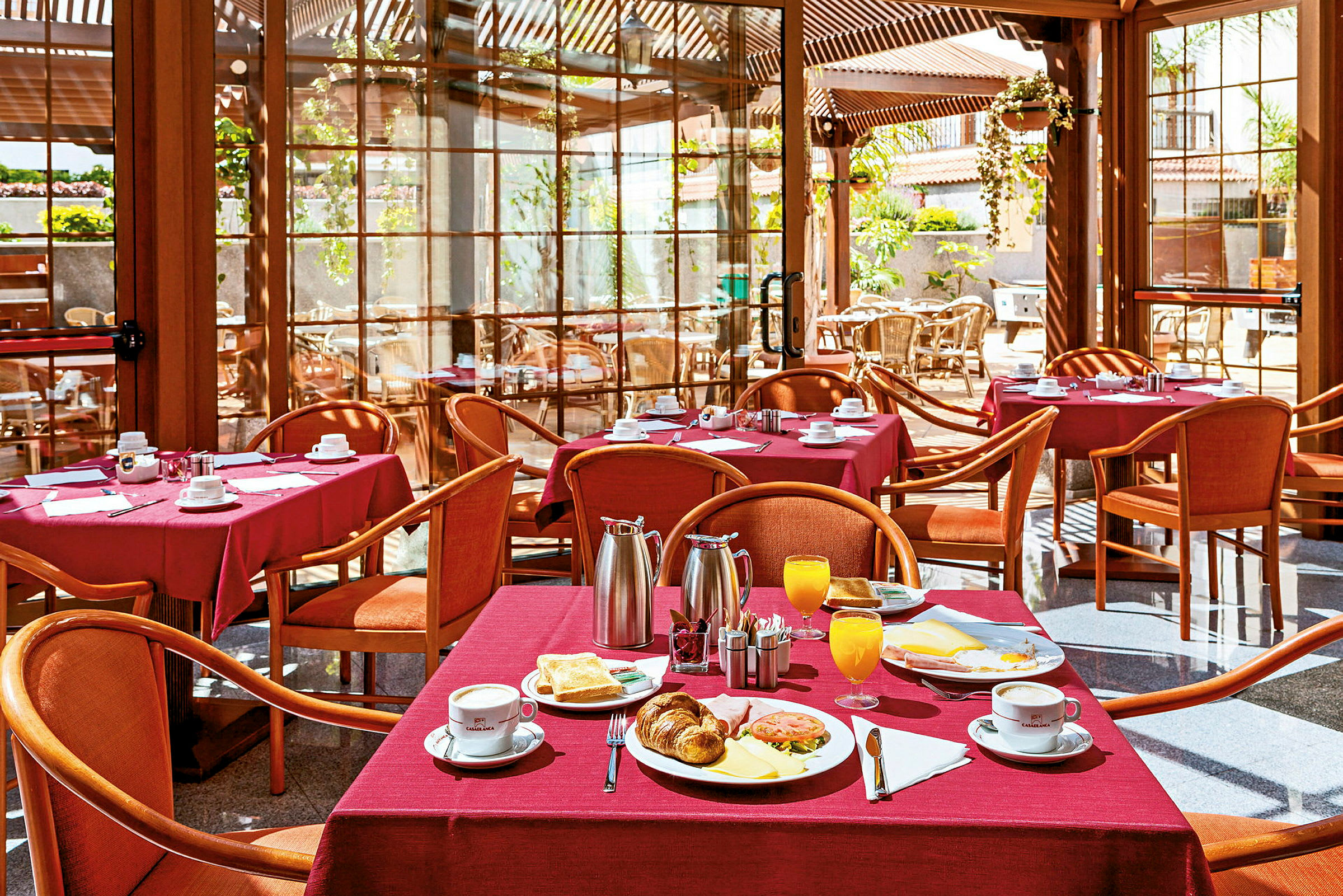 En restaurant med bord dekket med røde duker, serverer frokost inkludert kaffe, jus og bakverk. (AI-generert tekst)