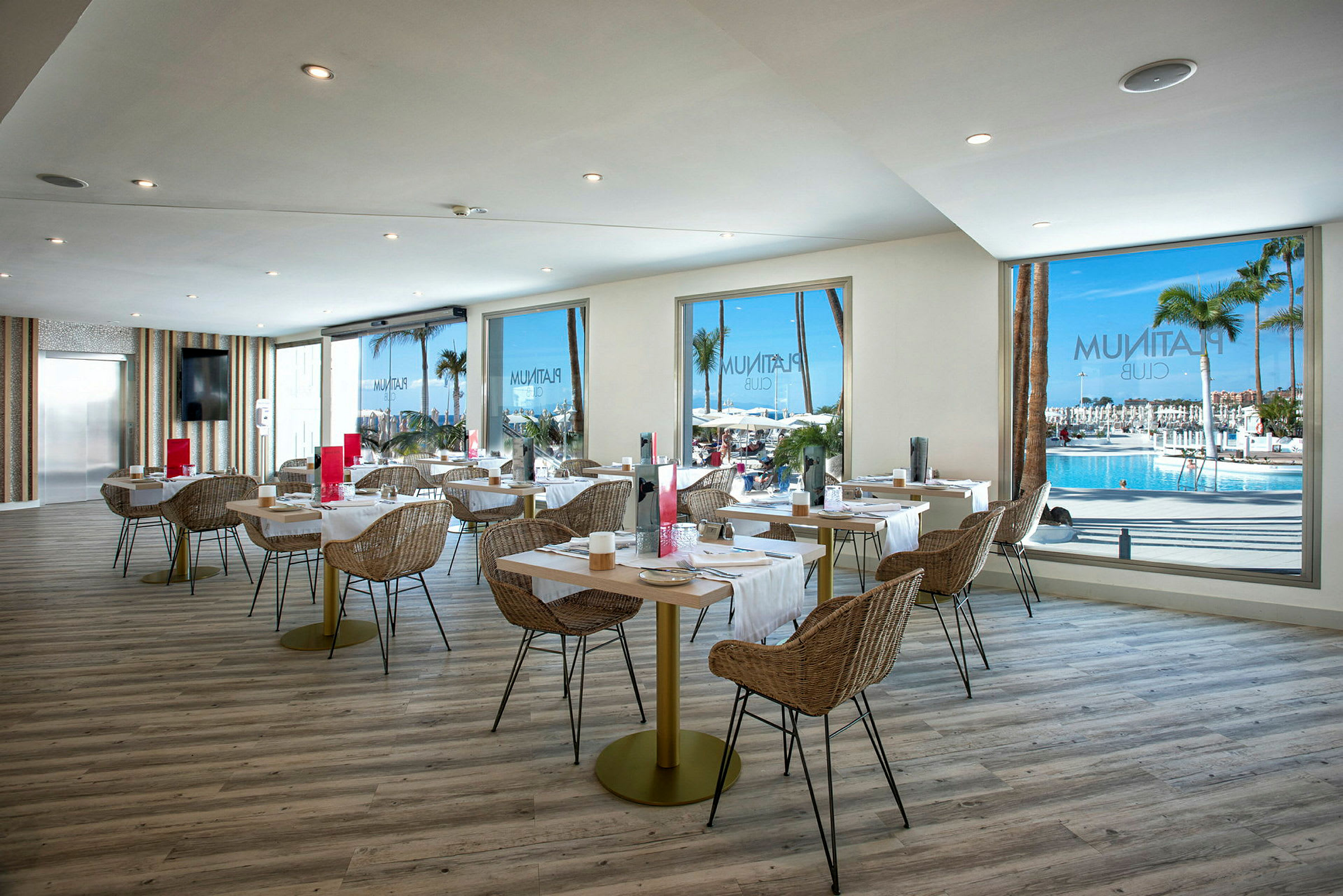 Innredning av en lys restaurant med utsikt mot et basseng og palmetrær gjennom store vinduer. (AI-generert tekst)
