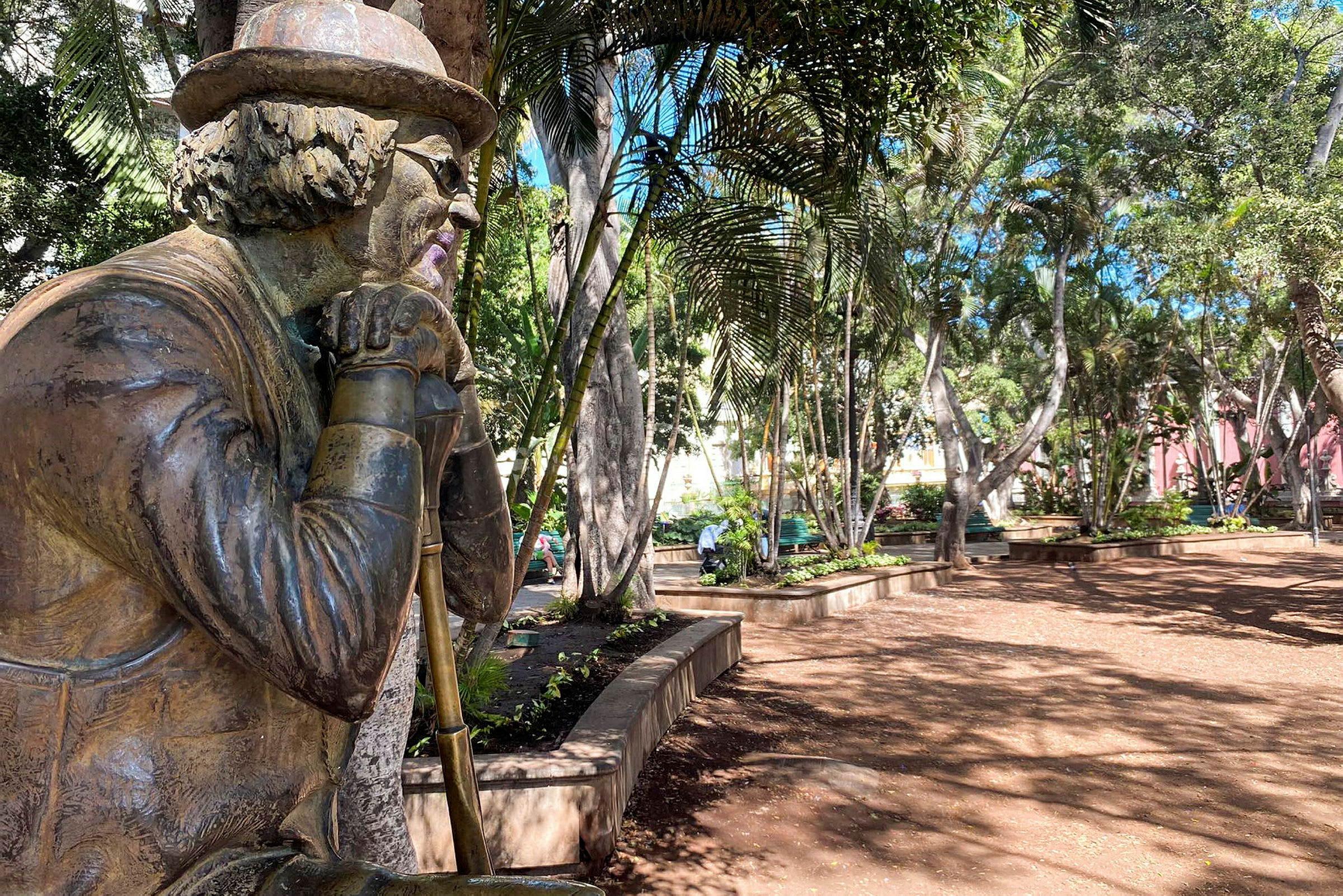 En statue av en person med hatt og briller, plassert i en frodig park. (AI-generert tekst)