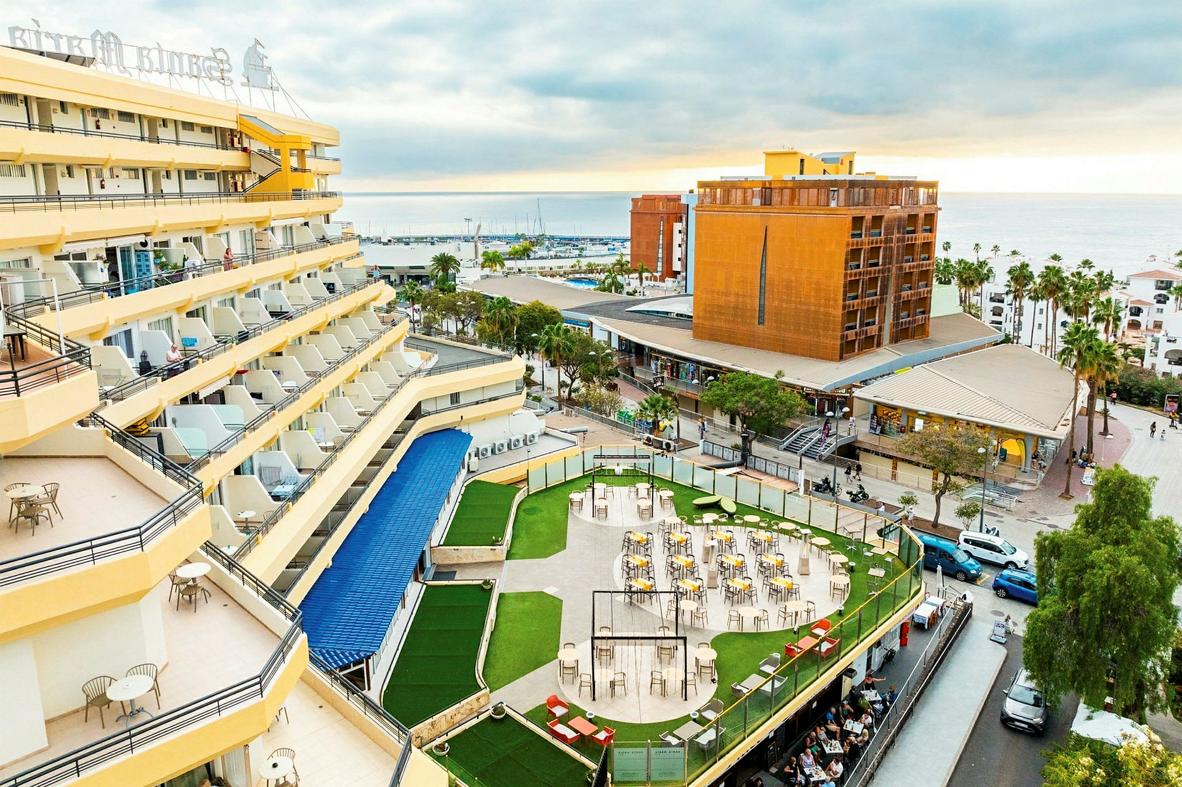 Hotell med gule fasader og balkonger, en terrasse med bord og stoler omgitt av grøntområder, og en utsikt over byen og havet i bakgrunnen. (AI-generert tekst)
