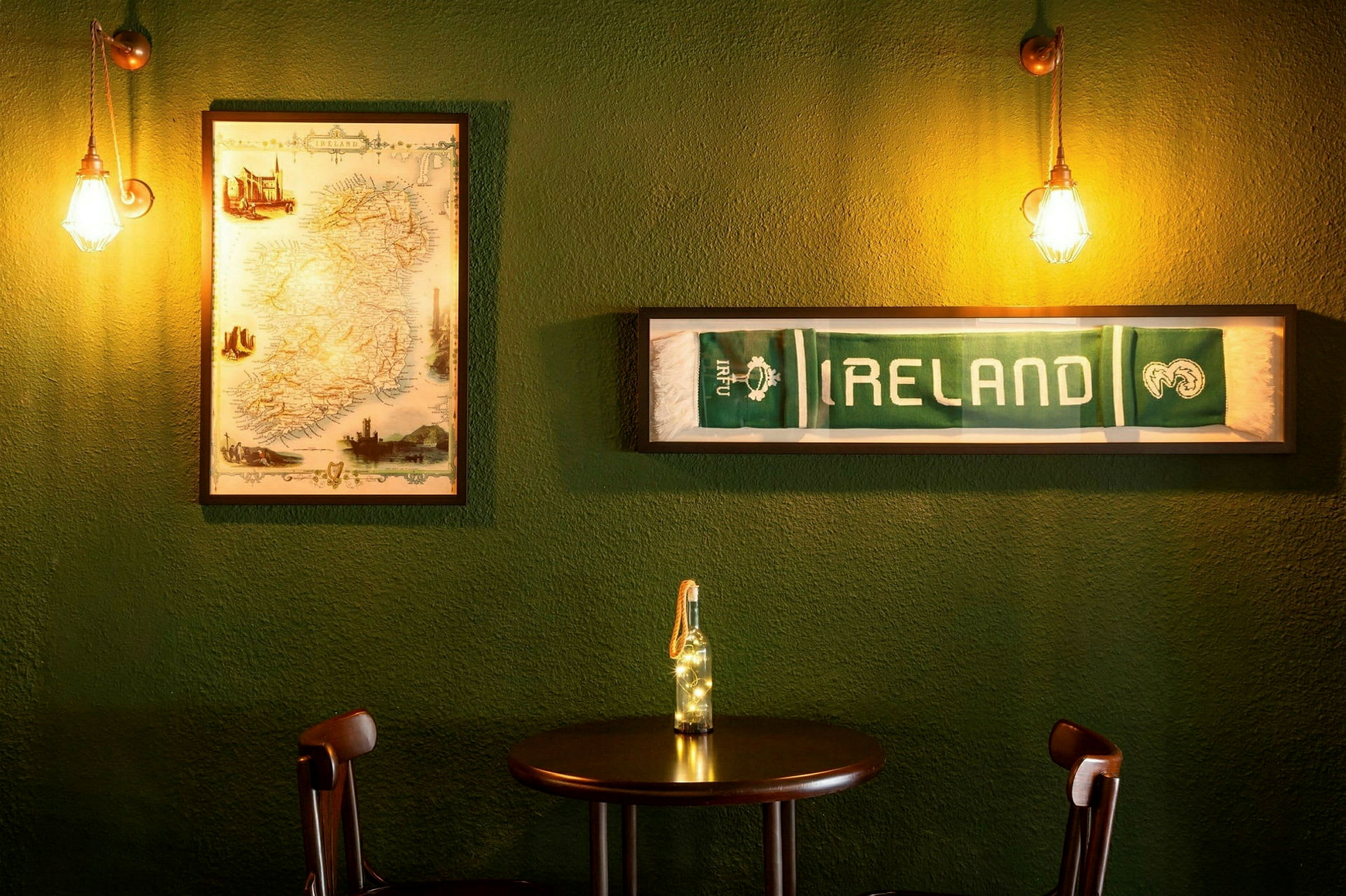 Et bord med to stoler foran en grønn vegg, dekorert med et innrammet kart over Irland og et innrammet skjerf med teksten "IRELAND". (AI-generert tekst)