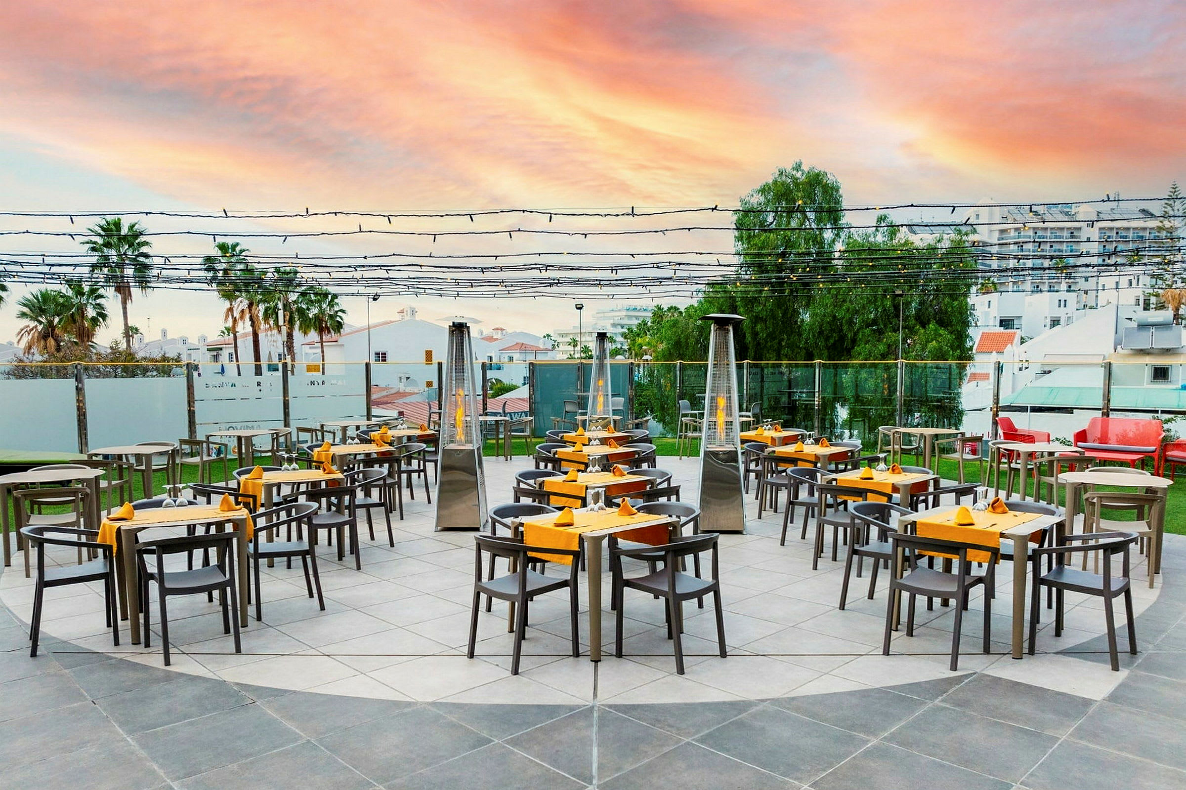 En utendørsrestaurant med bord og stoler arrangert på en steinlagt overflate under en himmel med solnedgangens farger. (AI-generert tekst)