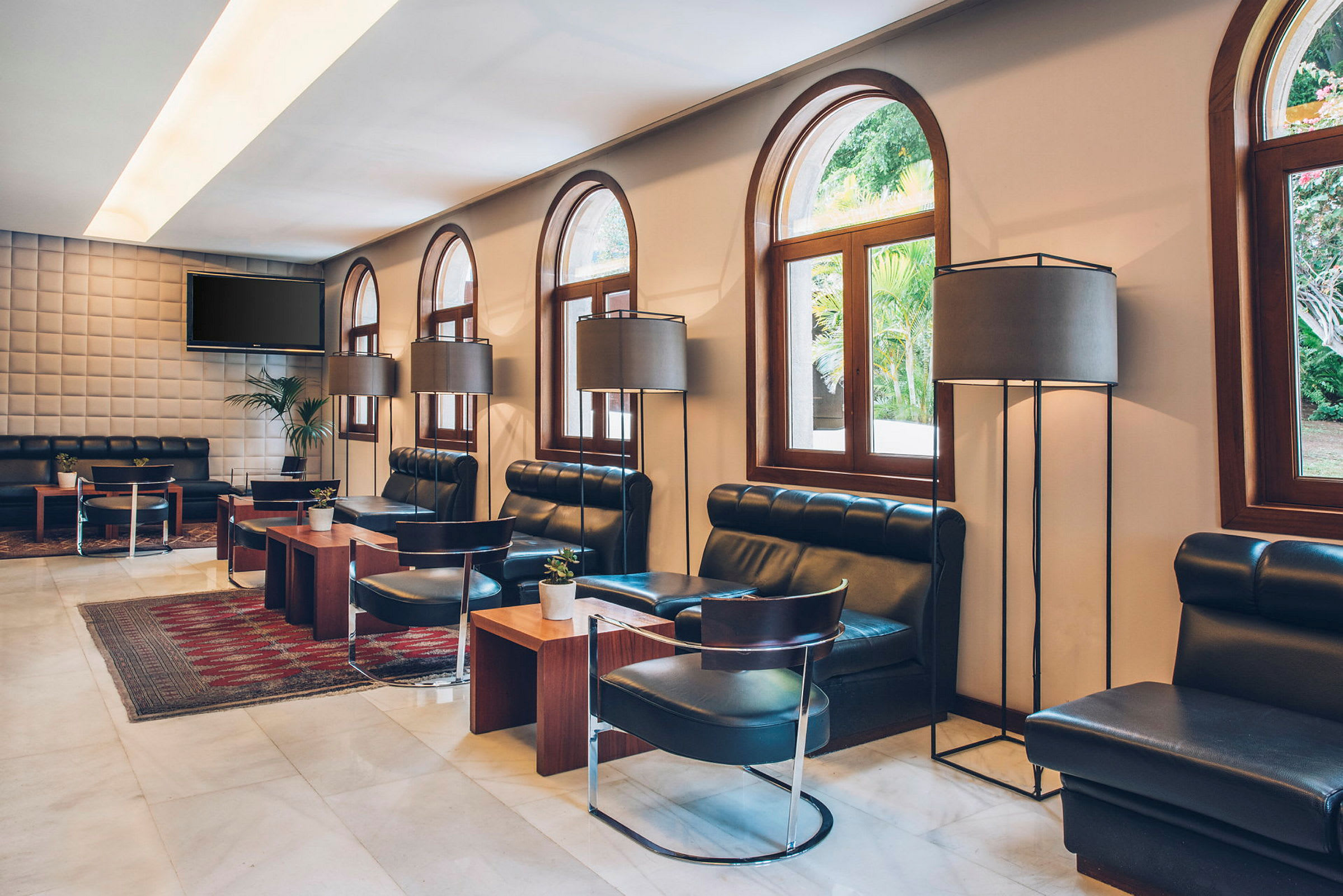 Et elegant loungeområde med mørke skinnsofaer, trebord og store gulvlamper. Runde vinduer gir utsikt mot grøntområde utenfor. (AI-generert tekst)
