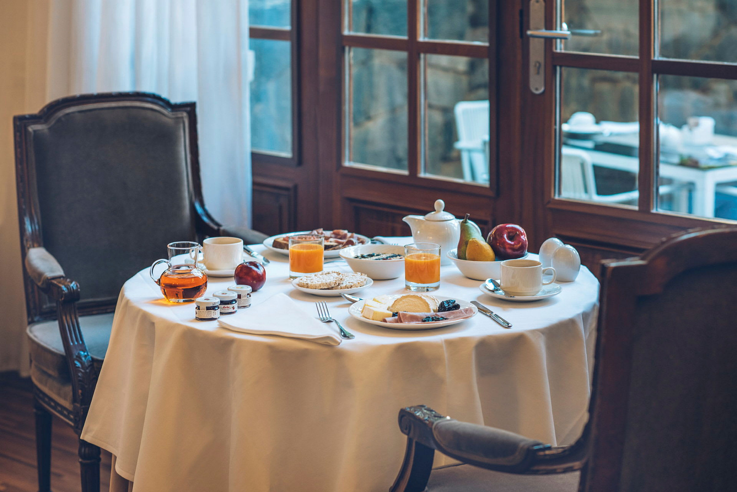 Et dekket frokostbord med glass appelsinjuice, te, frukt og oppskåret pålegg. Bordet står ved et vindu. (AI-generert tekst)