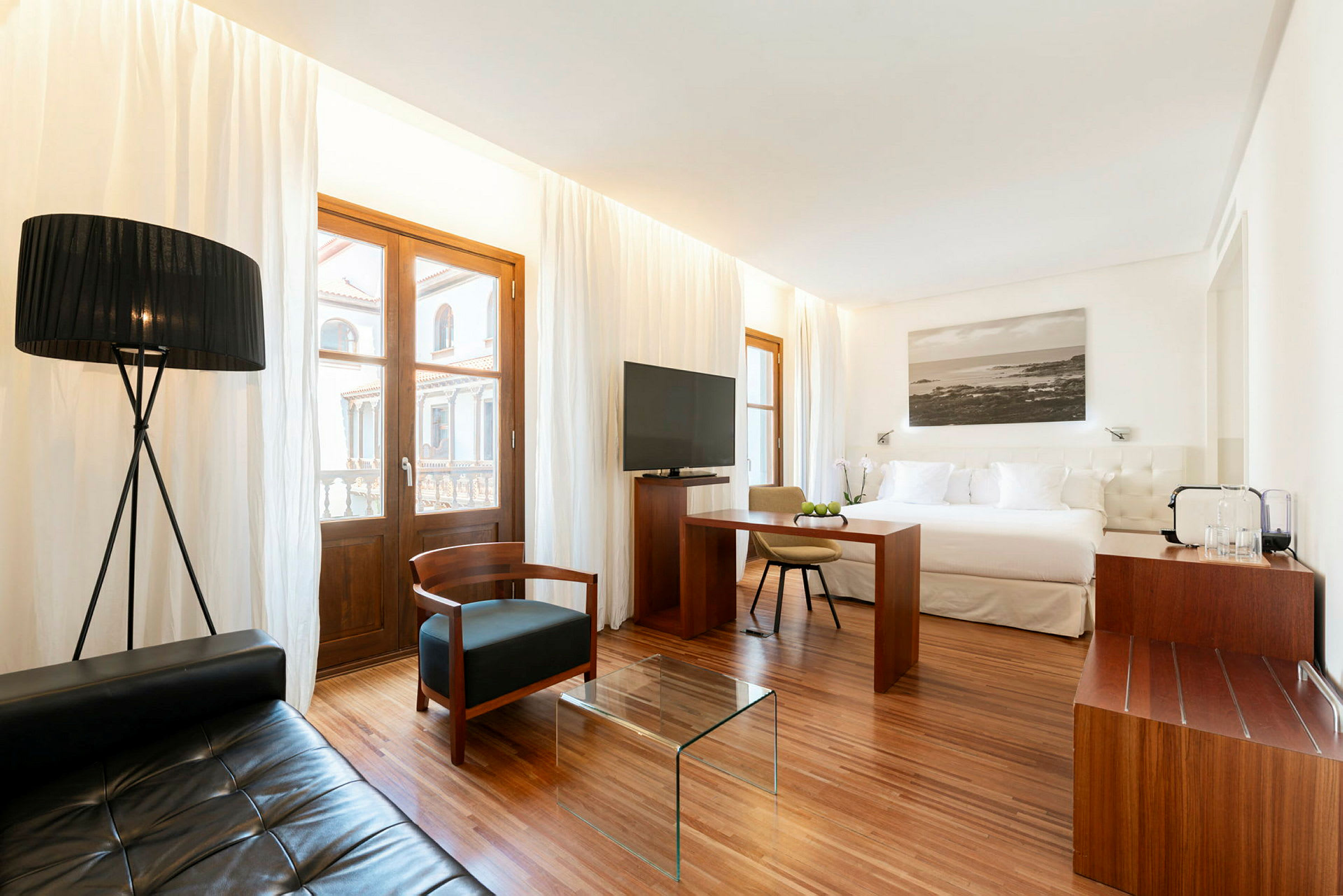 Et moderne innredet hotellrom med en stor seng, sittegruppe med tekstilmøbler og et glassbord, samt vinduer med hvite gardiner. (AI-generert tekst)