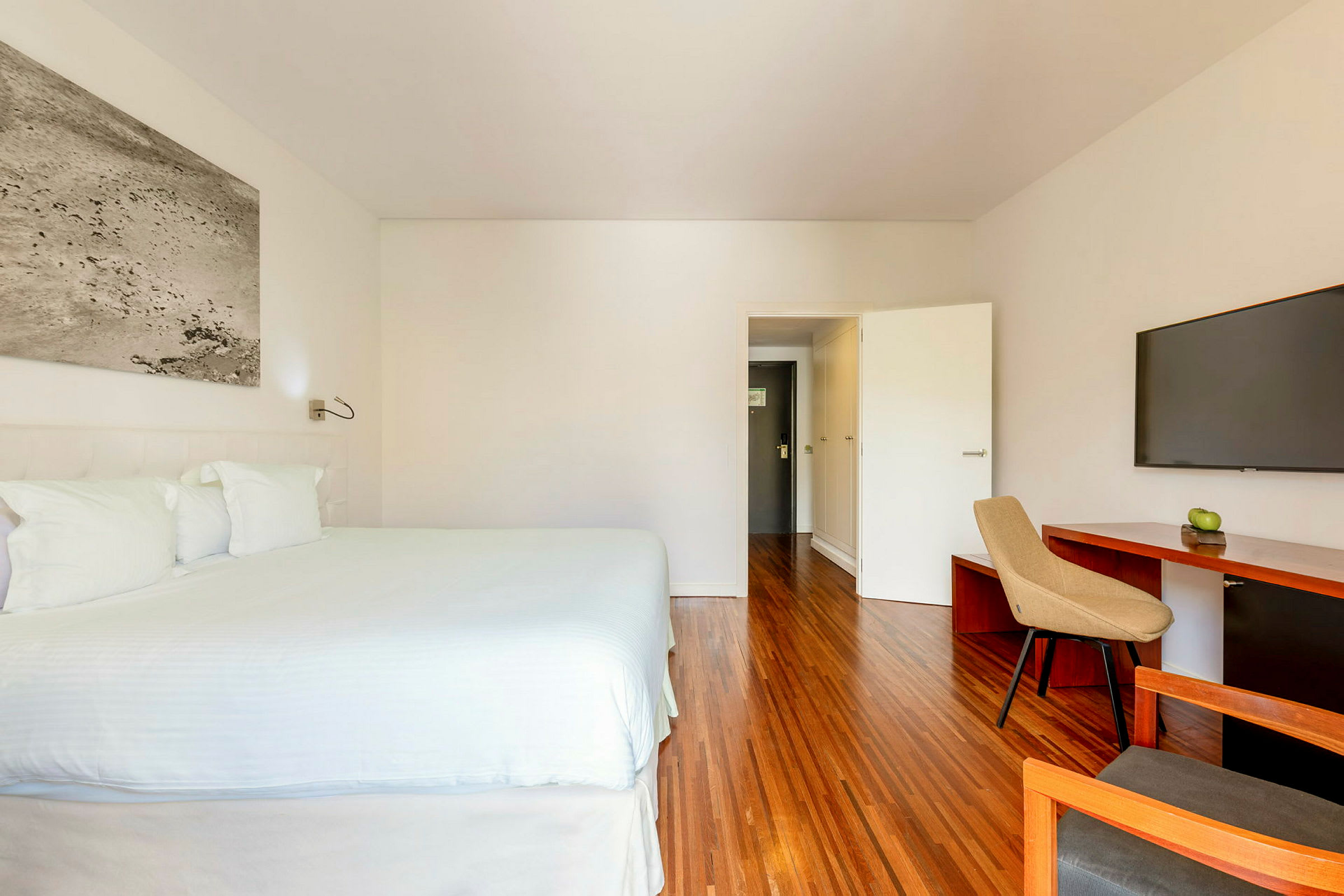 Et hotellrom med en stor seng ved siden av et sidebord, en TV på veggen og et skrivebord med en stol samt et eple på skrivebordet. (AI-generert tekst)