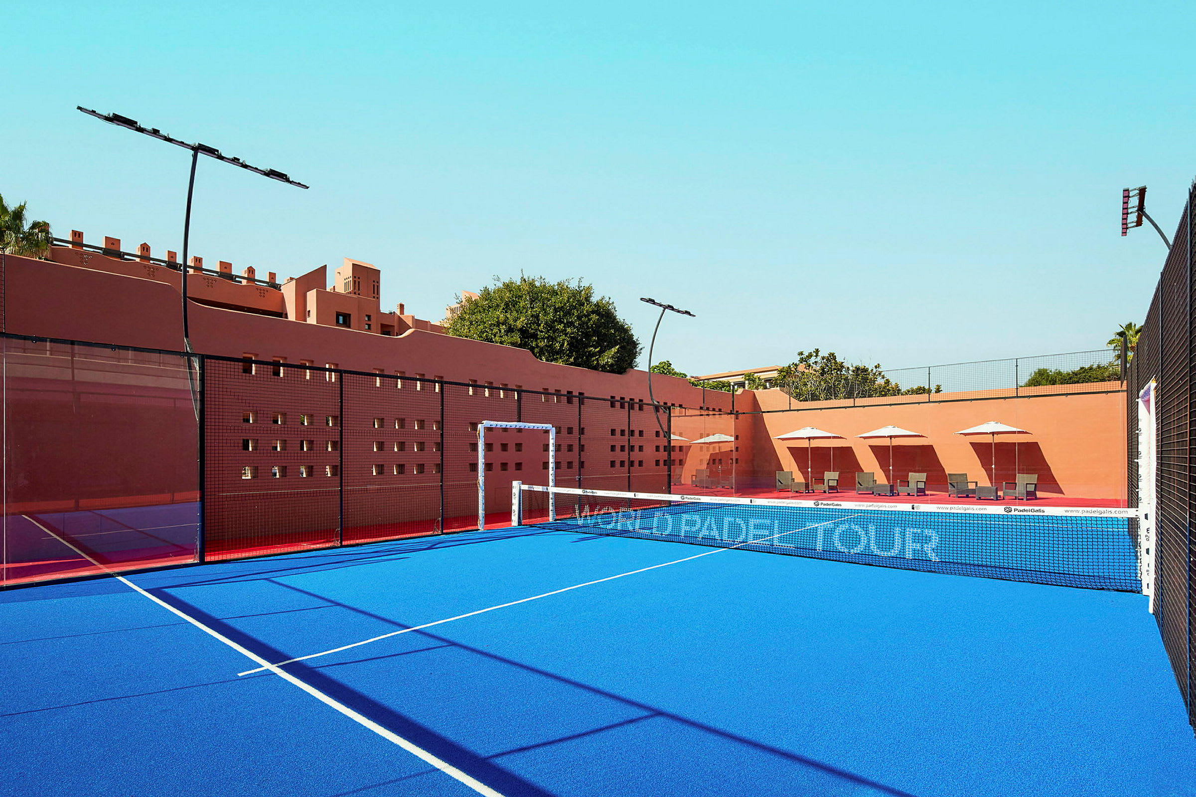 Padelbane med blått underlag omgitt av rosa vegger. Solstoler med parasoller vises i bakgrunnen. (AI-generert tekst)