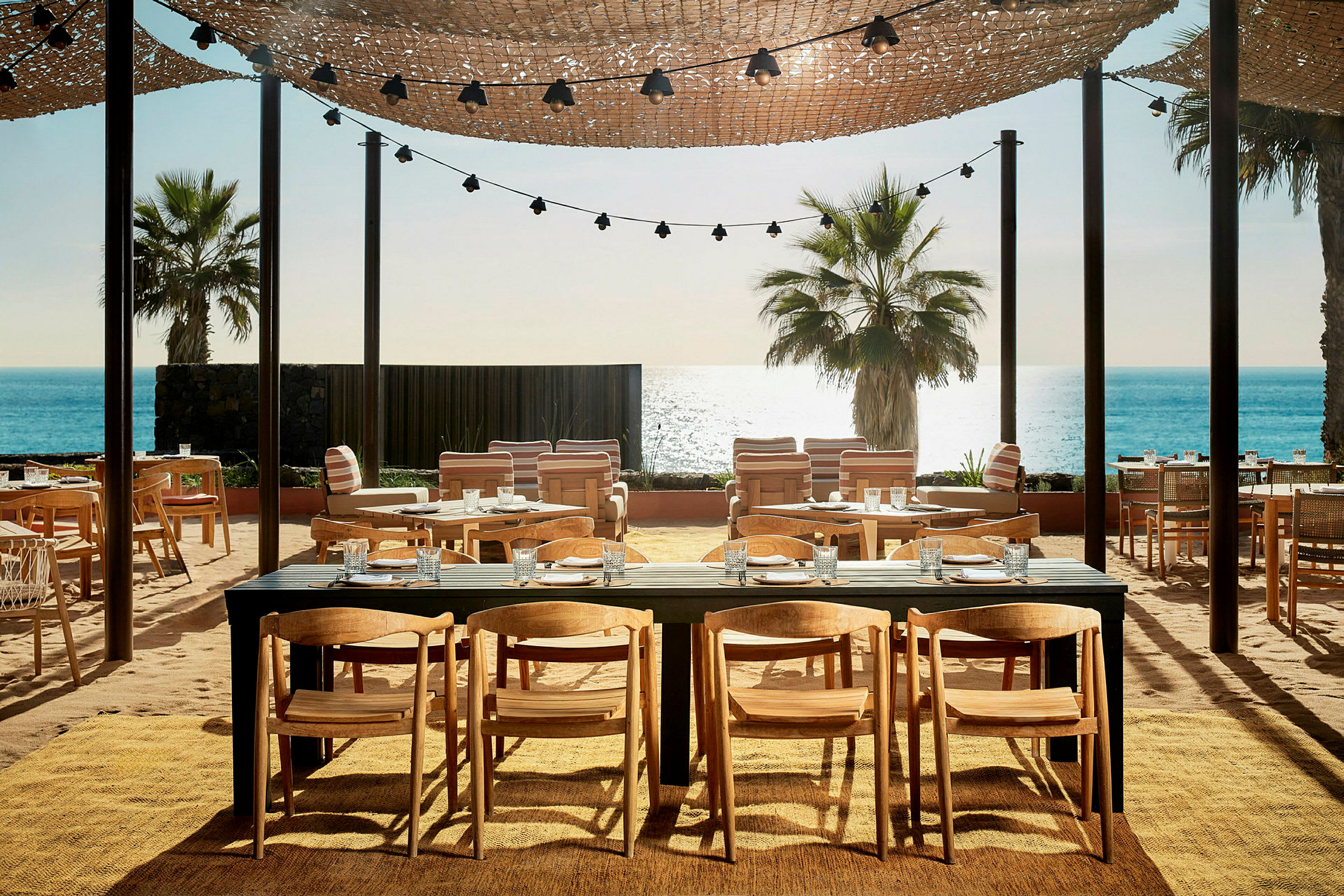Utendørs restaurant med bord og stoler, en palme og havet i bakgrunnen. (AI-generert tekst)