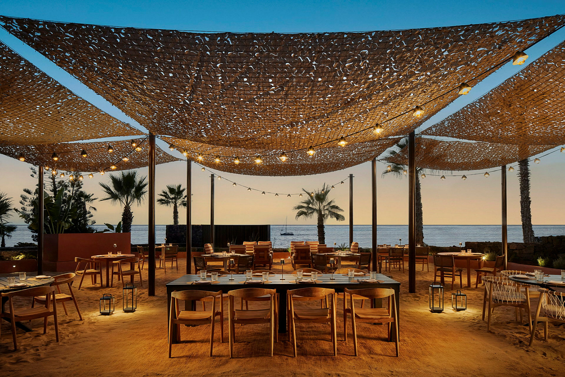 Utendørsrestaurant med havutsikt og bord plassert på sand, med trestoler og dekorative lysinstallasjoner. (AI-generert tekst)