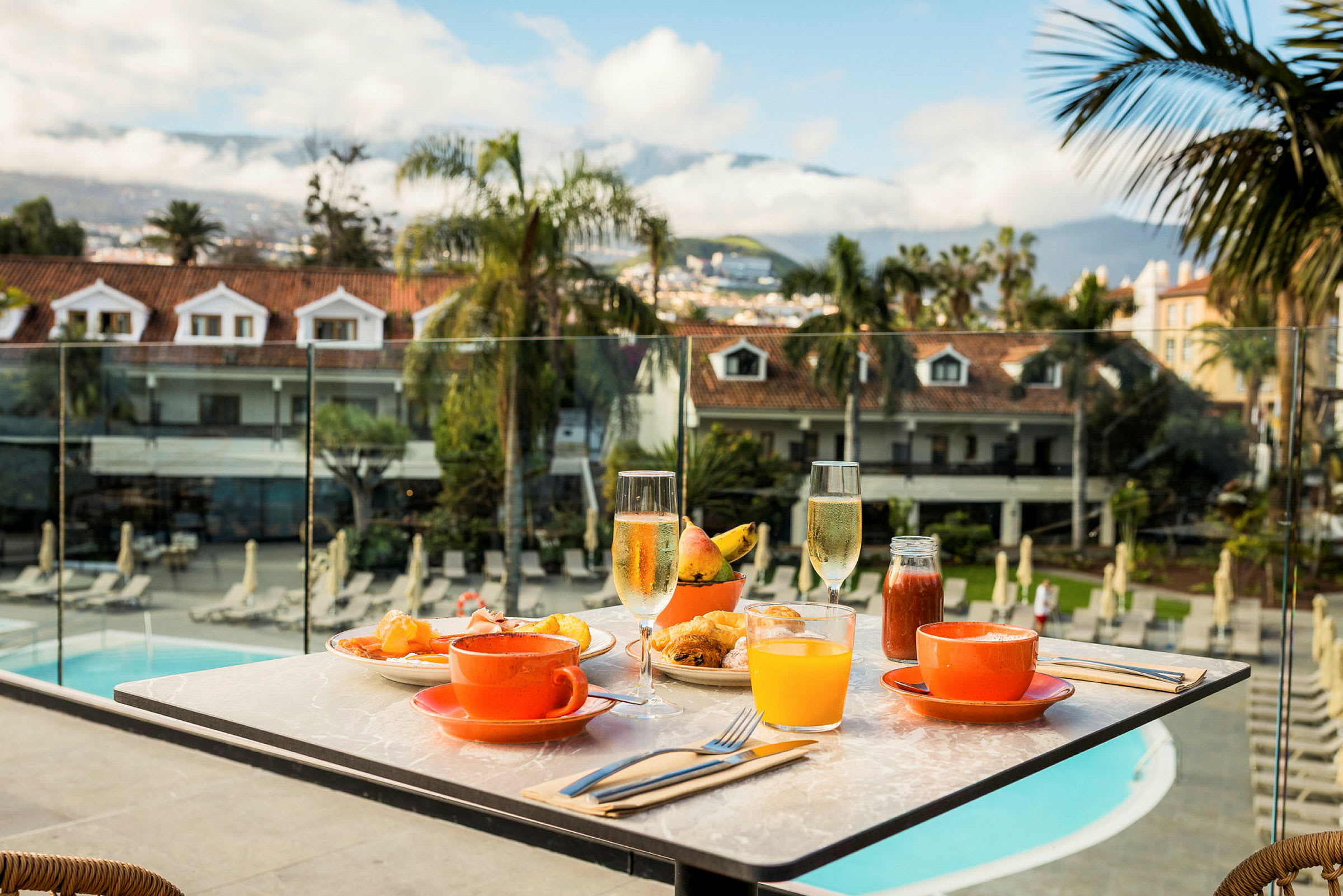Et dekket bord med frokostmat og drikke på en terrasse med utsikt over et bassengområde og omkringliggende natur. (AI-generert tekst)