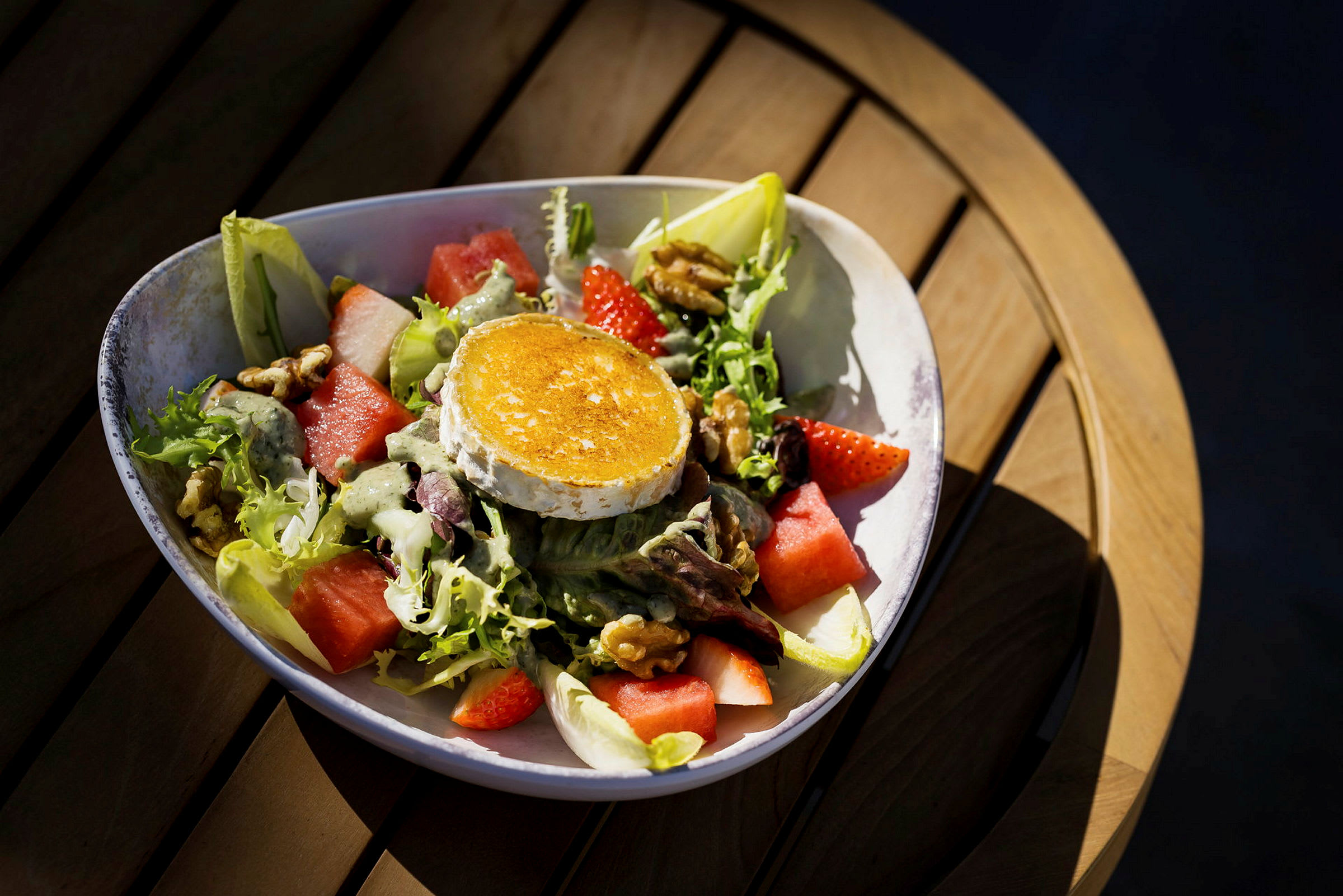 En tallerken med en salat som inneholder grillet osteskive, vannmelon, jordbær, nøtter og salatblader. Tallerkenen står på et trebord. (AI-generert tekst)