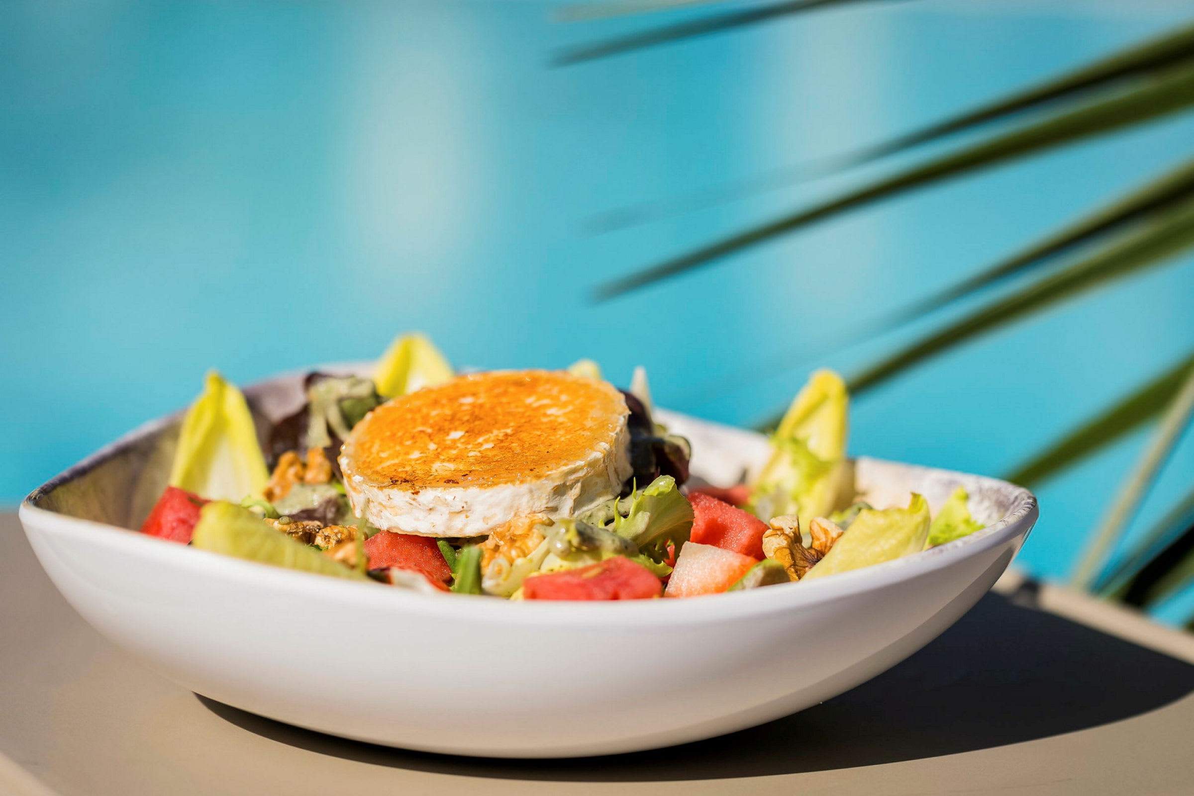 En salat med forskjellige grønnsaker og et stykke gratinert ost servert i en hvit bolle, med uskarp bakgrunn av blått vann og grønne blader. (AI-generert tekst)