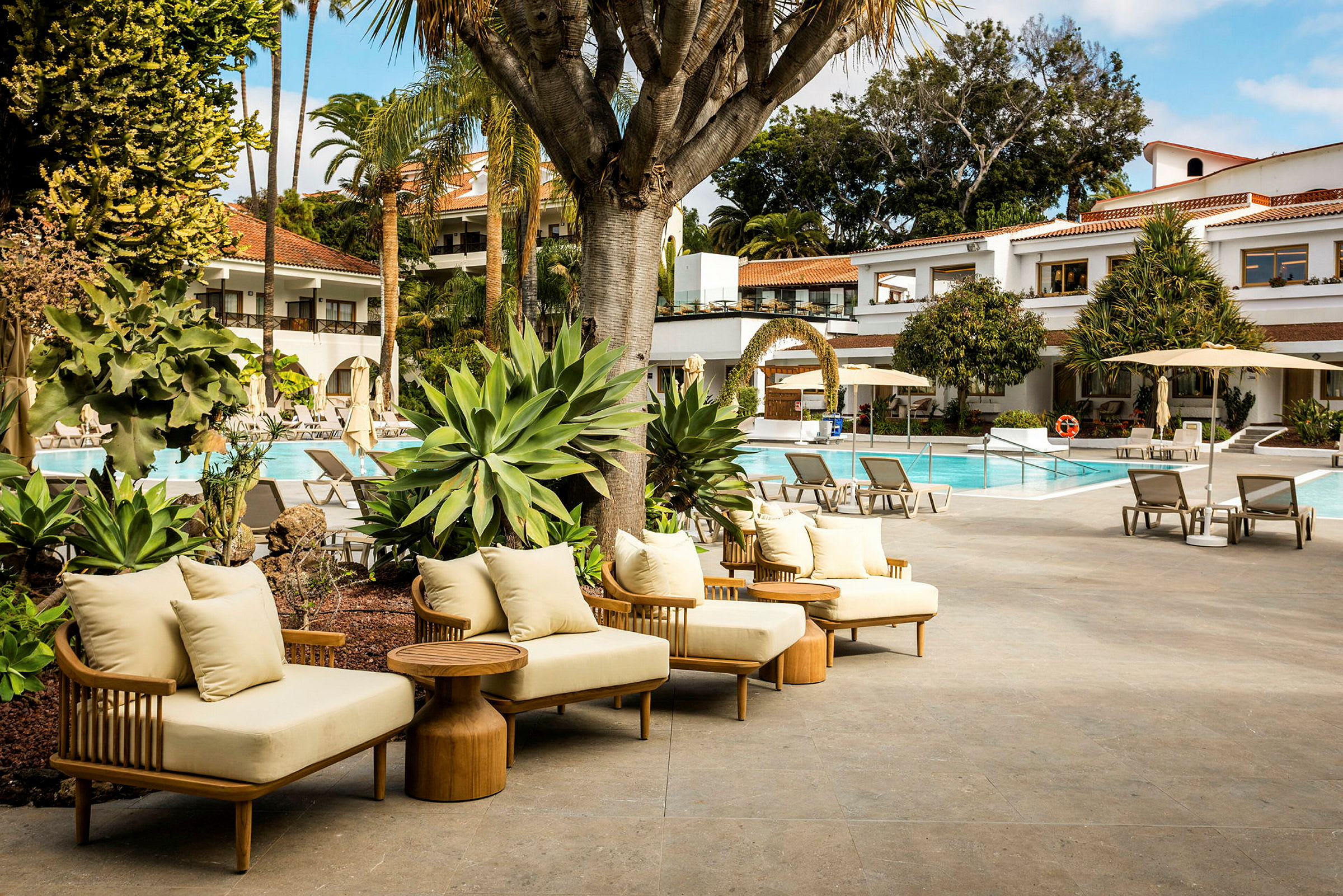 Et svømmebasseng med solsenger og parasoller omgitt av grønne planter og bygninger i bakgrunnen. (AI-generert tekst)