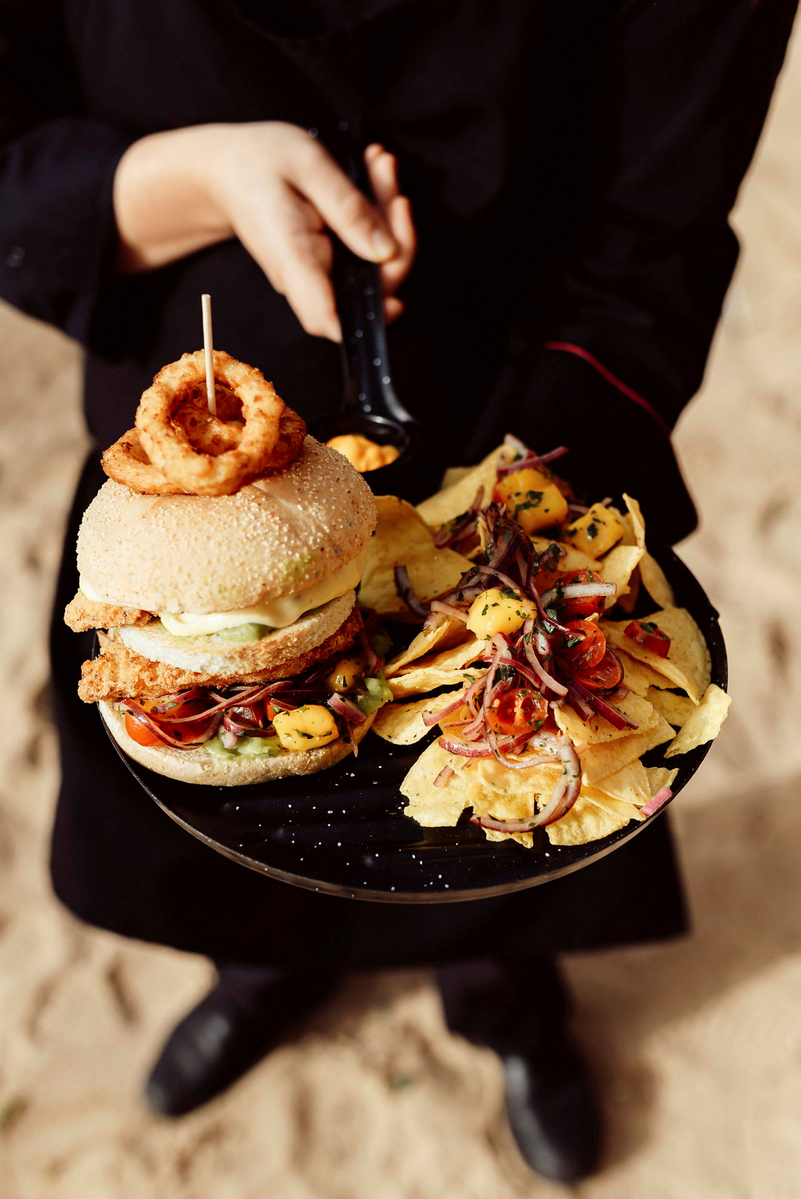 En person holder en tallerken med en burger toppet med løkringer og en side av nachos med salat. (AI-generert tekst)