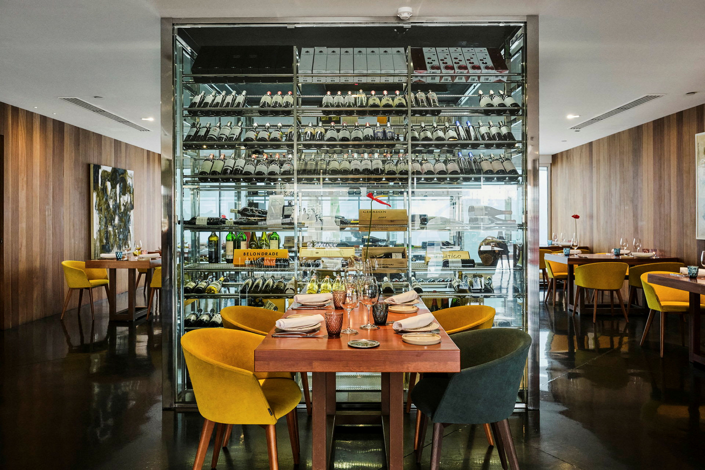Restaurant med et bord klargjort for gjester foran en glassvegg med lagrede vinflasker. (AI-generert tekst)