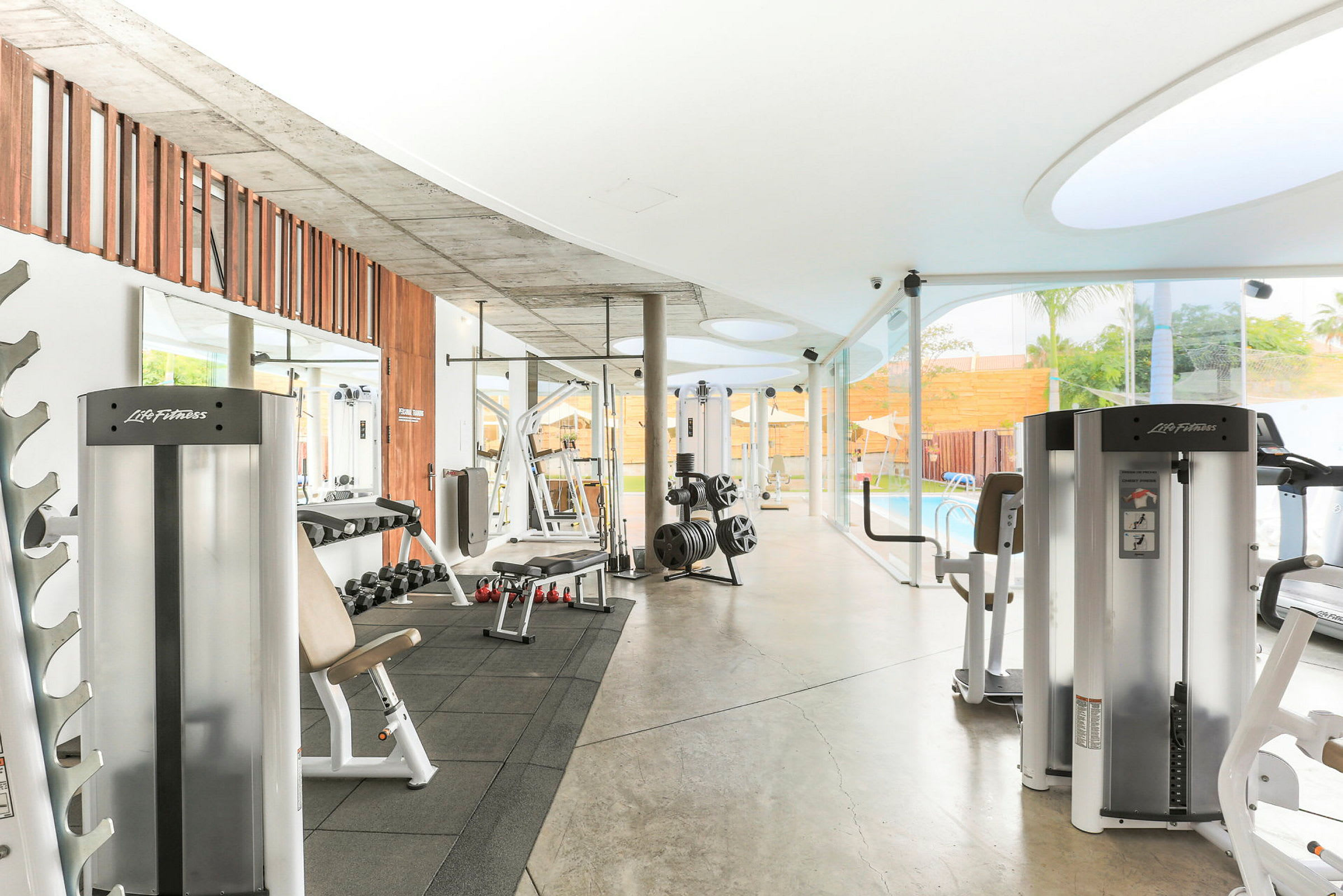 Et moderne treningsstudio med treningsapparater og frivekter, plassert nær en glassvegg med utsikt over et basseng. (AI-generert tekst)