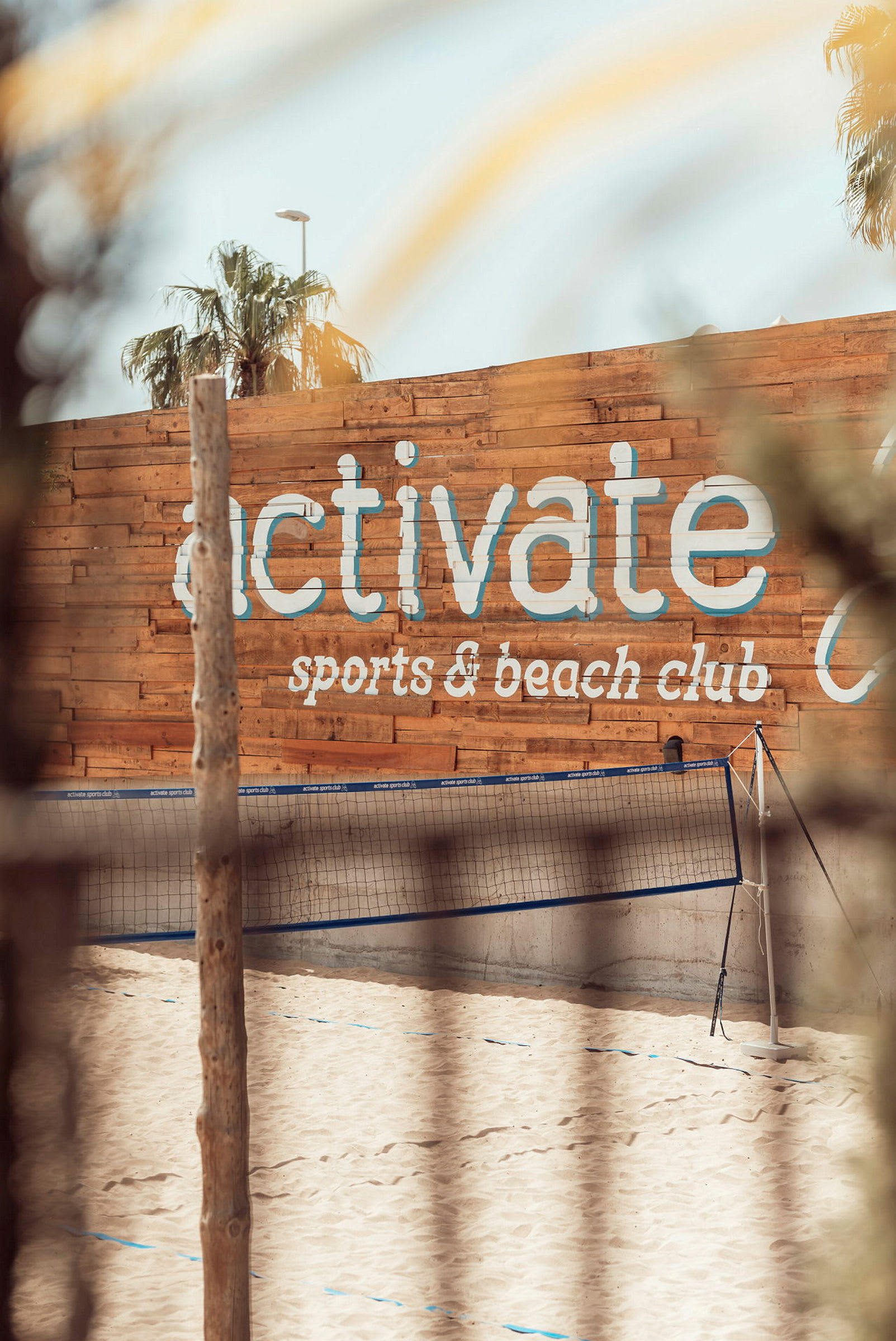 Sandstrand med volleyballnett foran en trevegg med teksten 'activate sports & beach club'. (AI-generert tekst)
