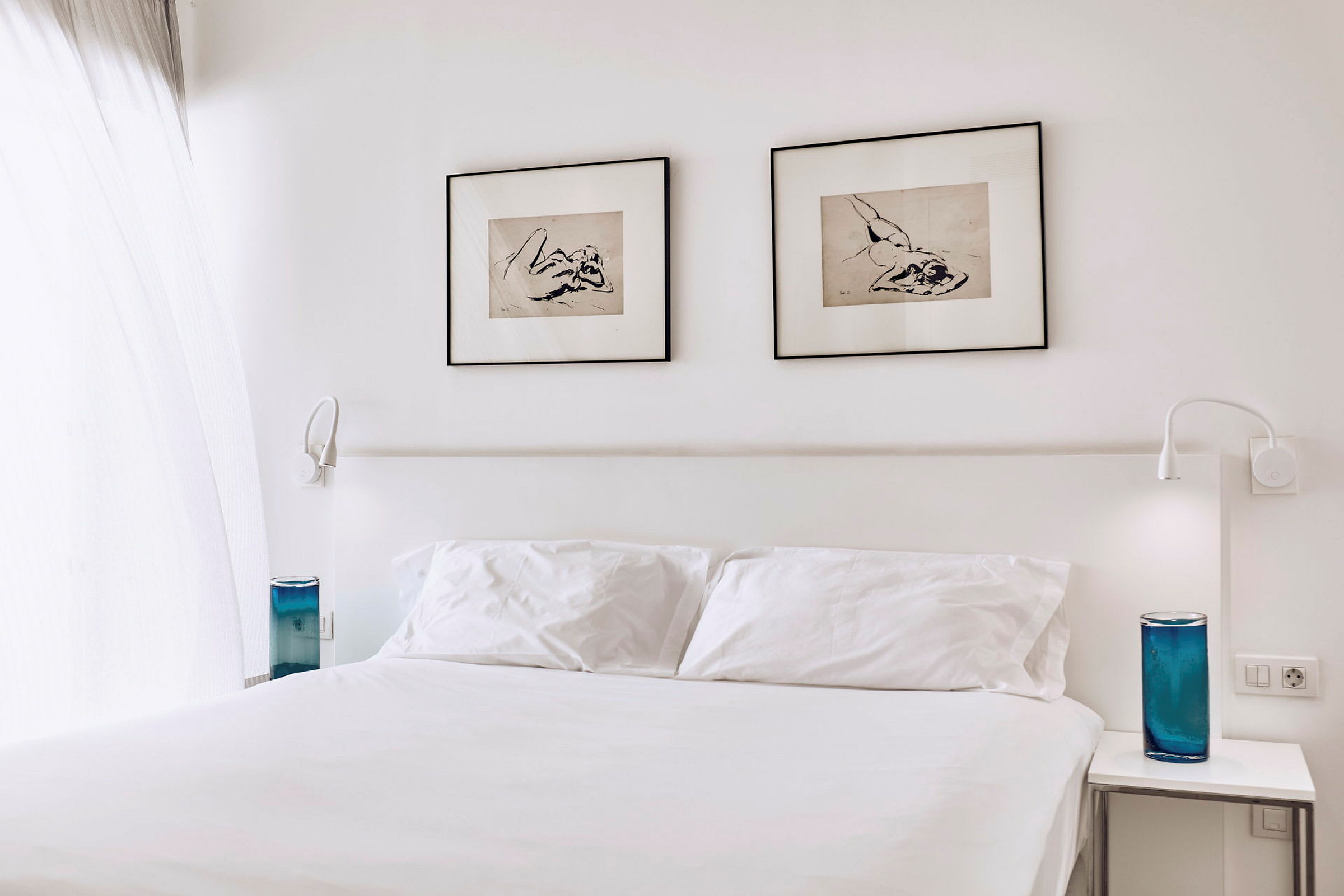 Et hotellrom med en hvit dobbeltseng, to innrammede kunstverk på veggen og blå dekorative gjenstander på nattbordene. (AI-generert tekst)